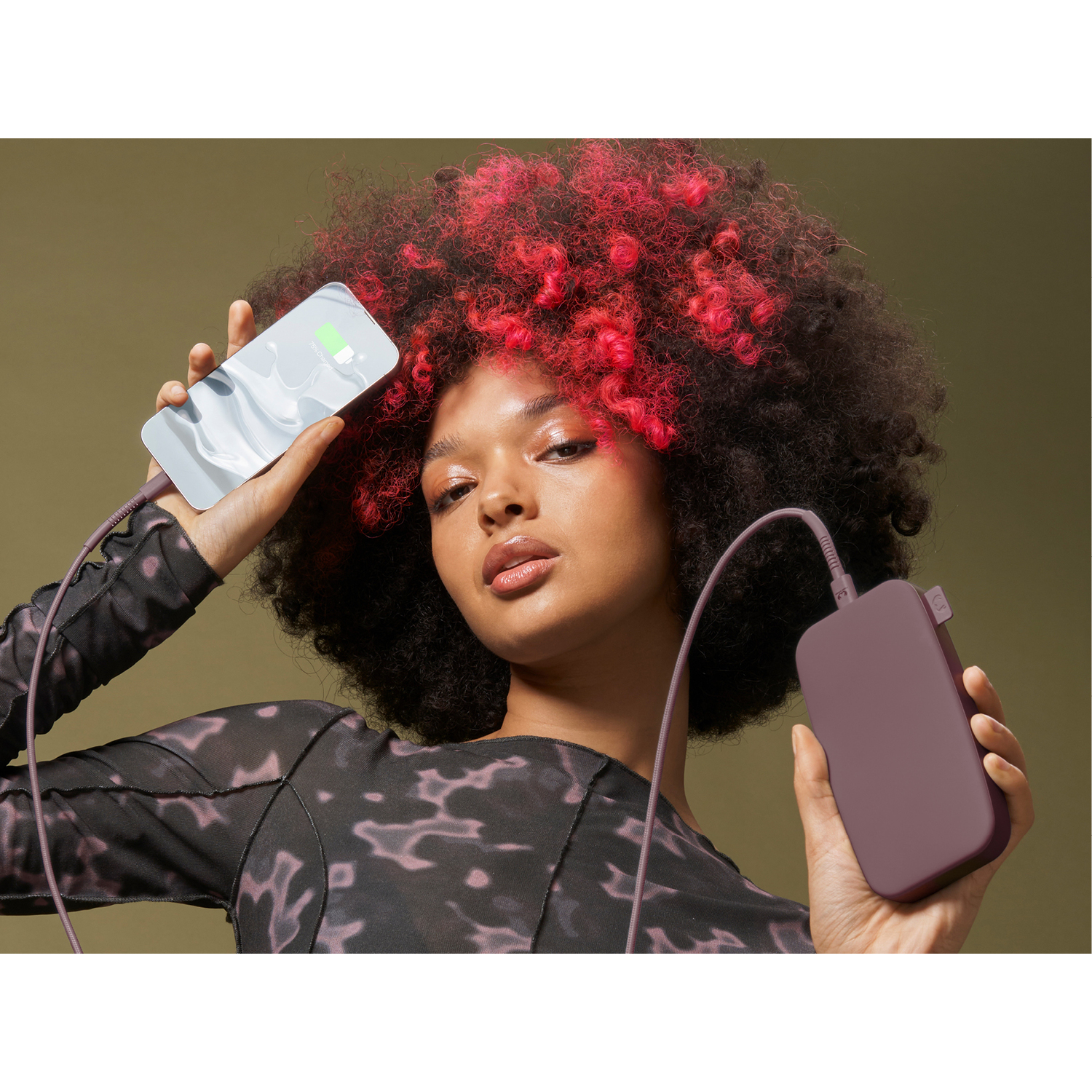 Une femme aux cheveux rouges tient un téléphone qui se charge à partir d'une batterie externe. Le fond est vert olive.