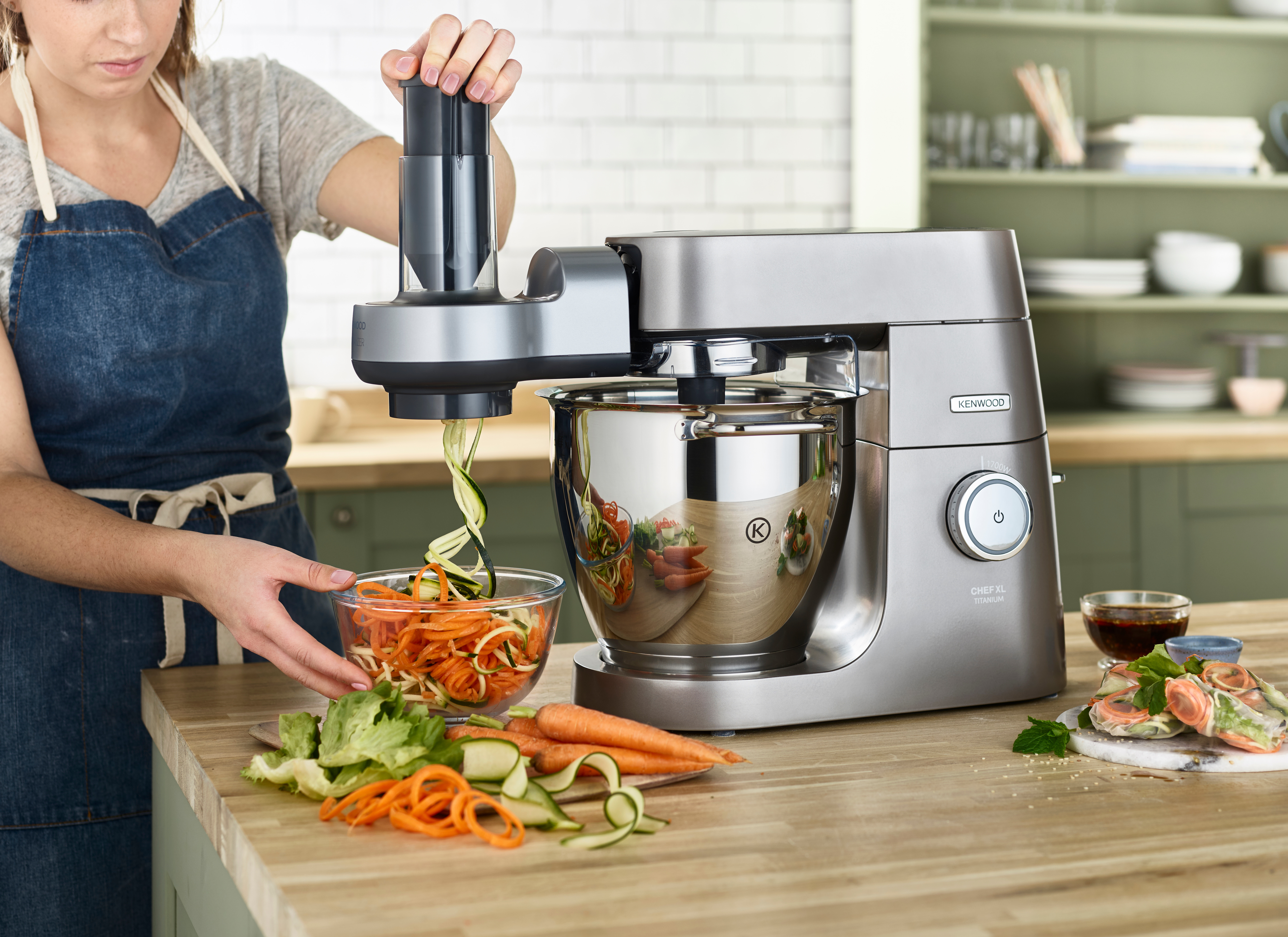 Frau benutzt silbernen Küchenmixer, bereitet Essen mit Gemüse auf einer Holztheke zu.