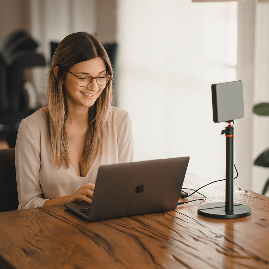 Femme avec des lunettes souriante, utilisant un ordinateur portable sur une table en bois. Lampe à droite.