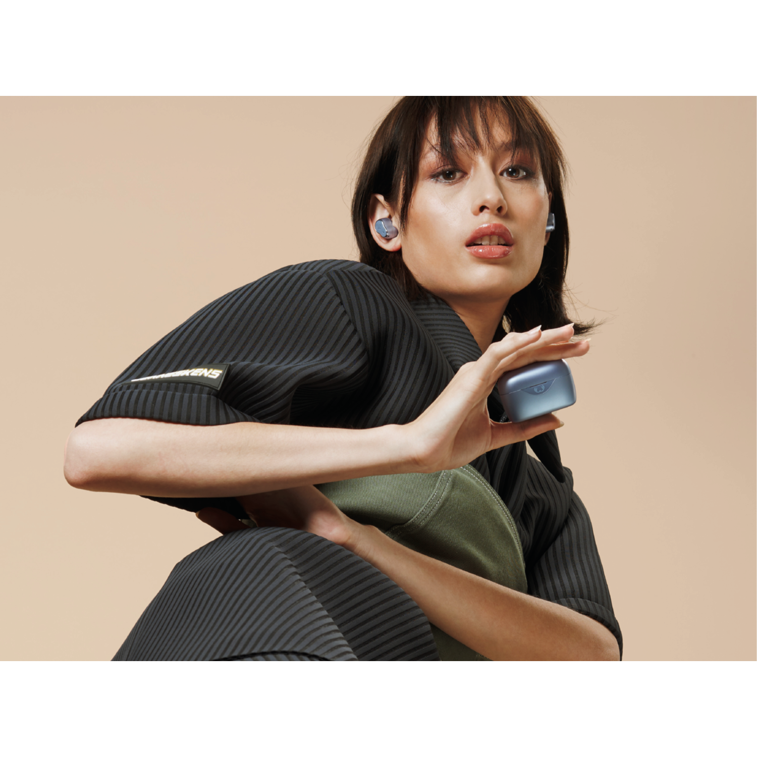 Woman holds a blue earbud case. Her black top and olive green bag contrast the beige background.