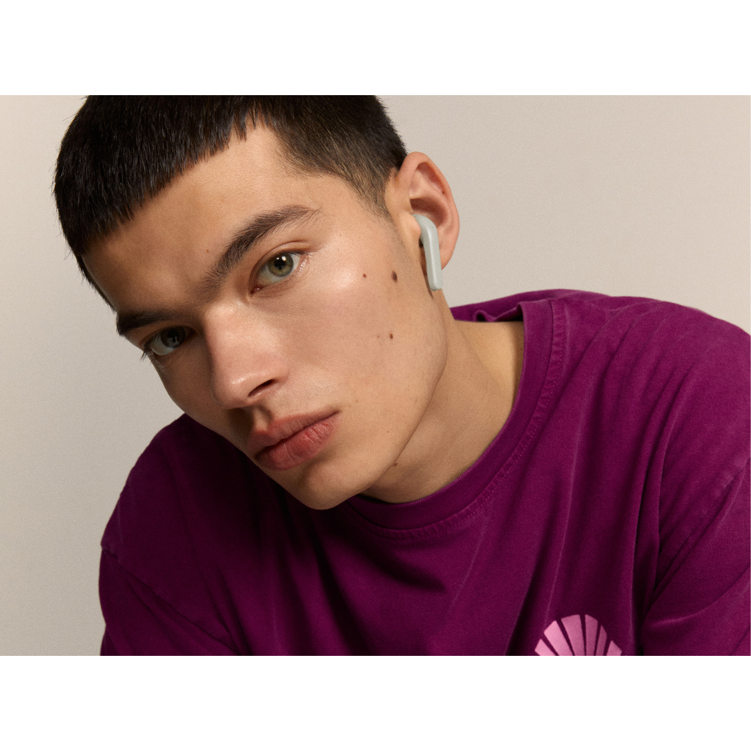 A young man with an earbud in his ear, looking towards the camera. Light background.