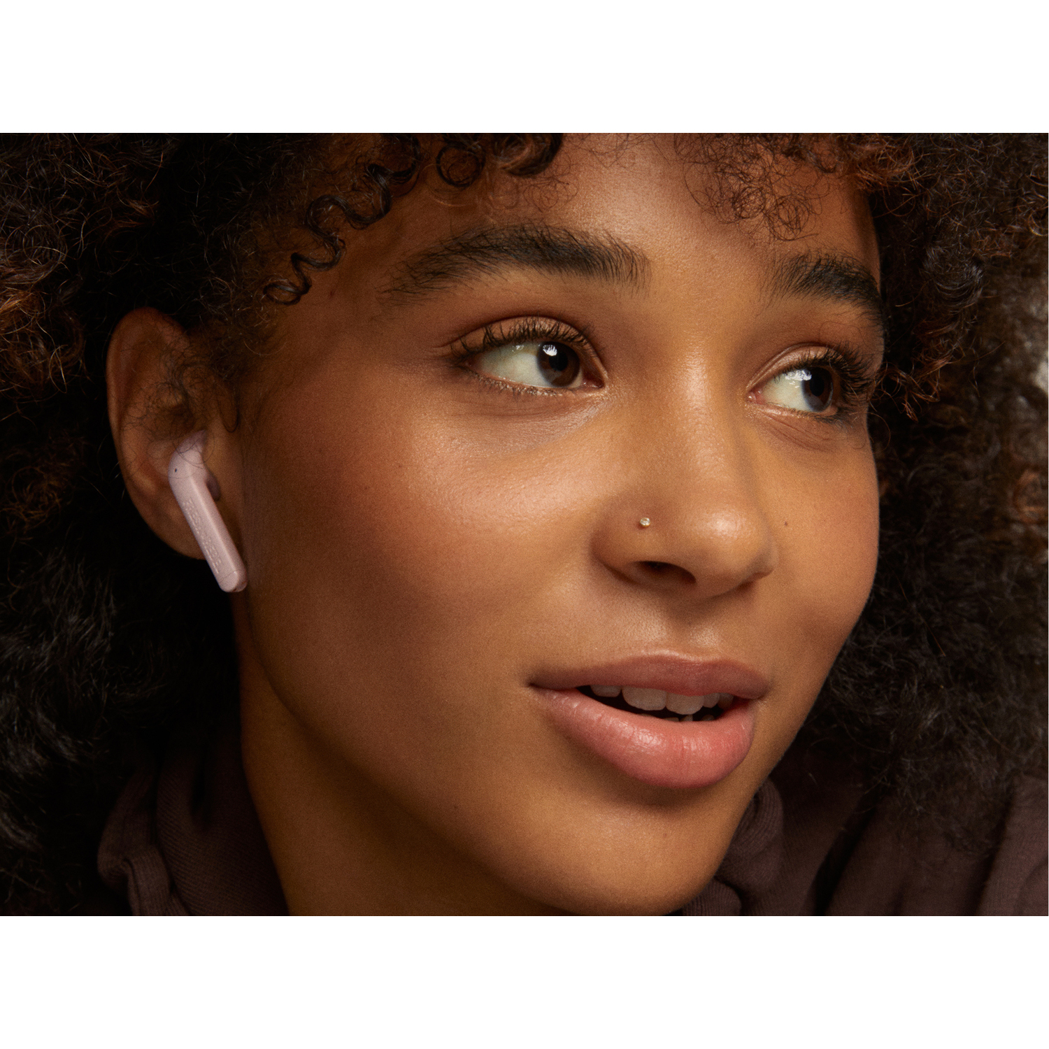 Une femme à la peau foncée lève les yeux, portant un écouteur. Elle a un piercing au nez.