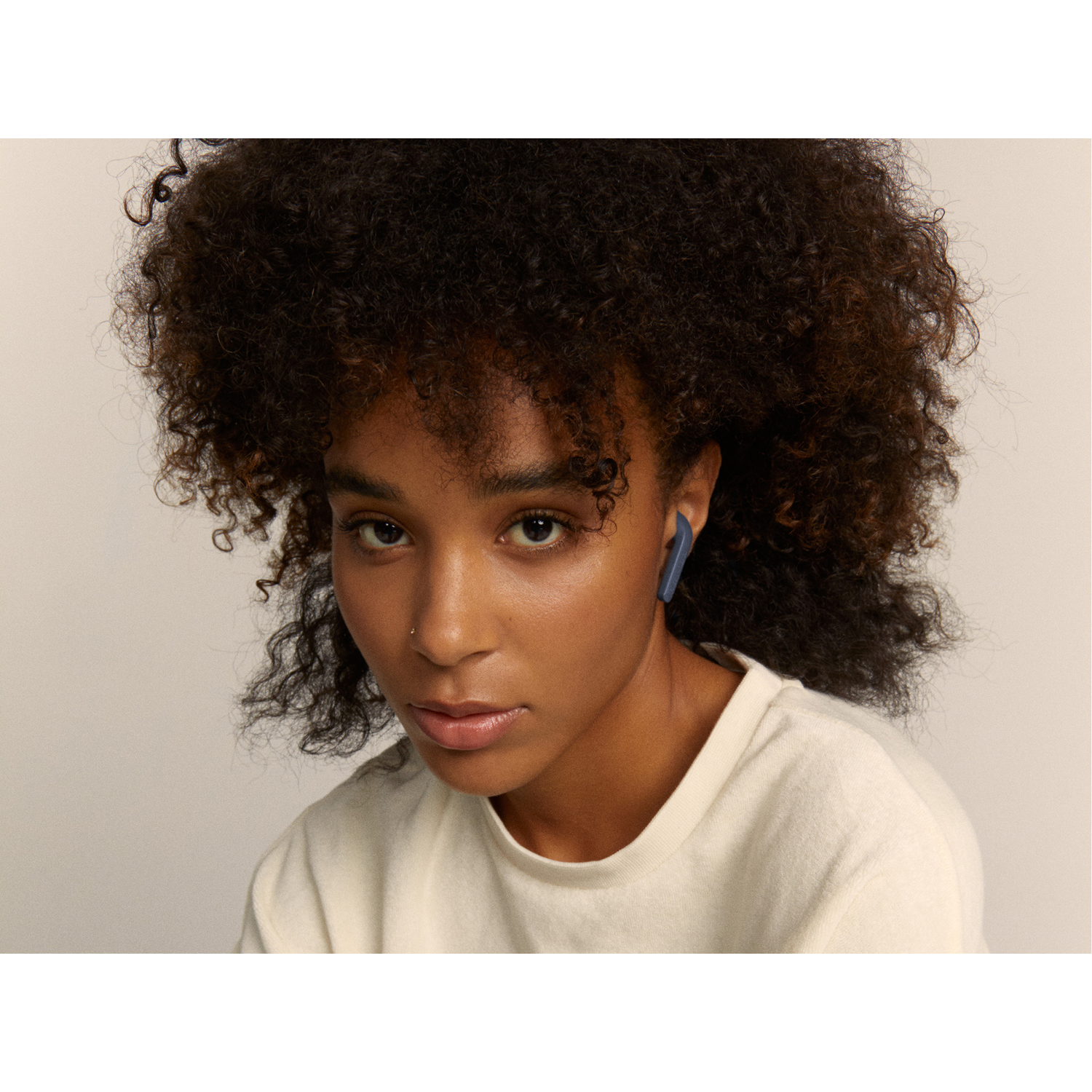 Young woman with curly hair wearing a white shirt, blue earbud in ear, looking at the camera.