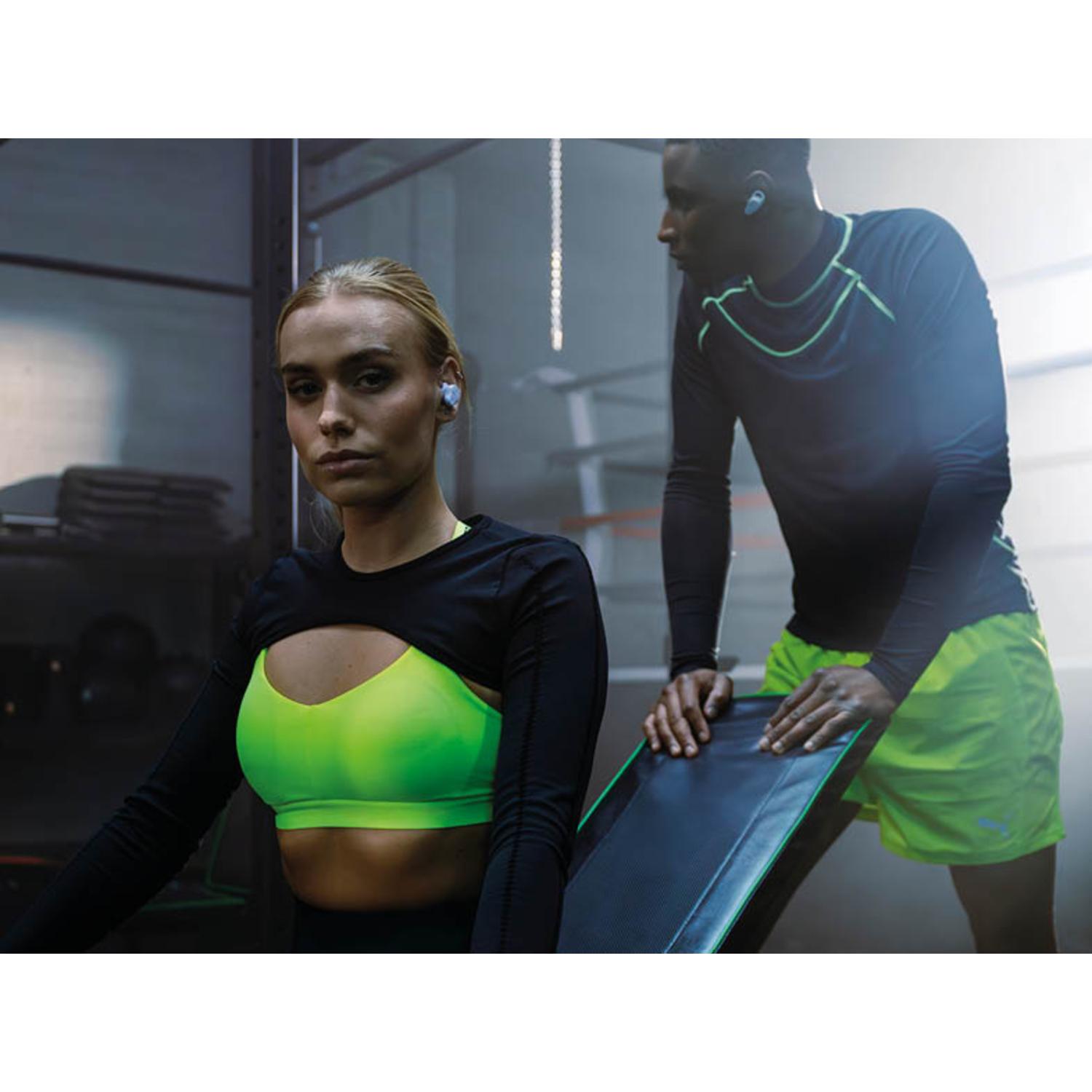 Man and woman in gym with earbuds. Green and black outfits.