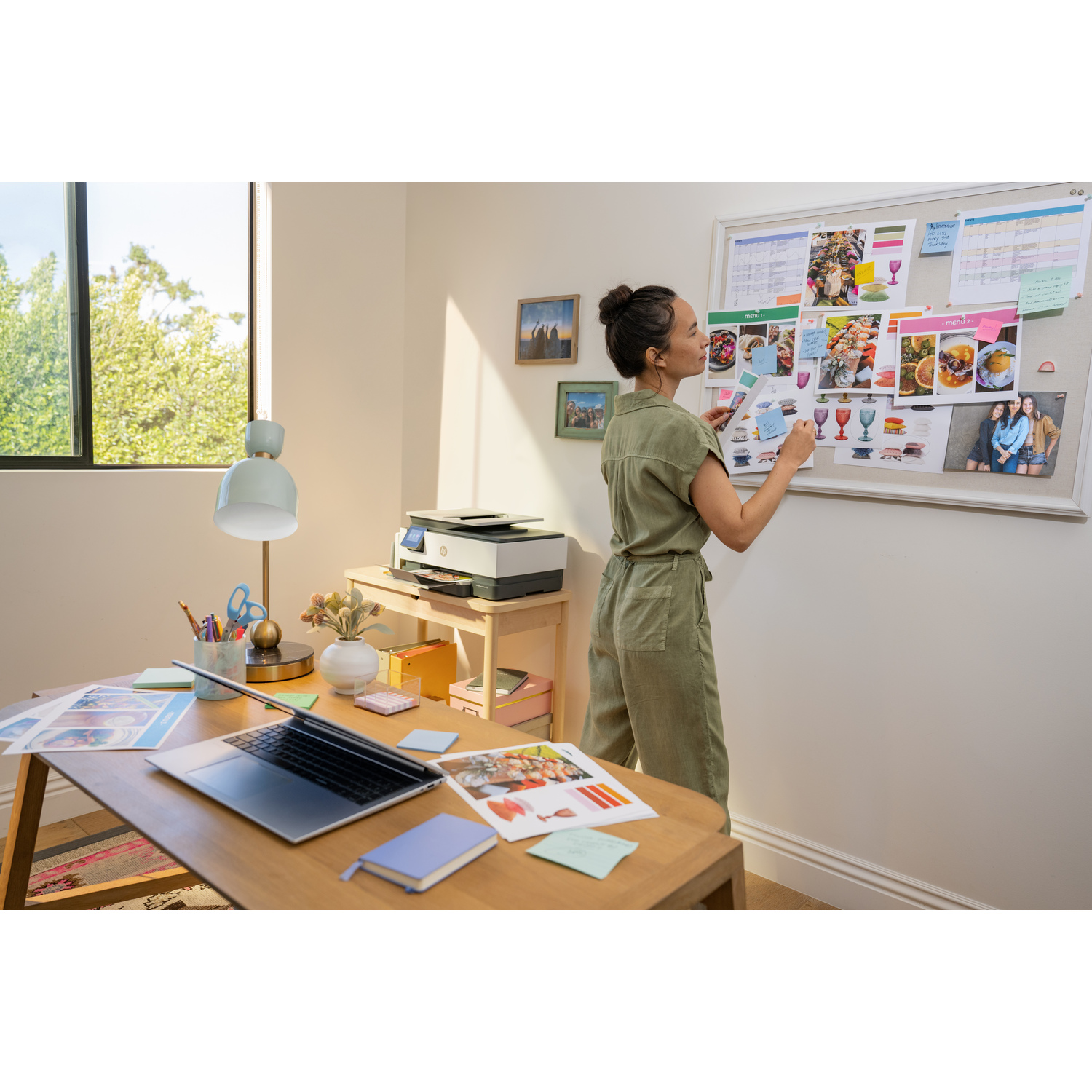 A woman works at a desk with a laptop, printer, and papers, arranging notes on a board.