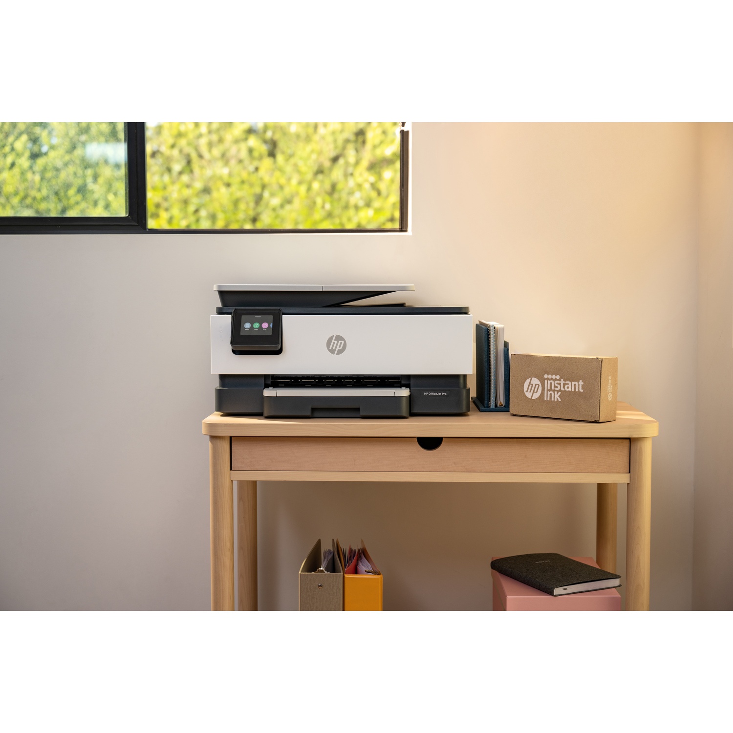 A printer on a wooden desk with a box of instant ink. Books and a notebook are also present.