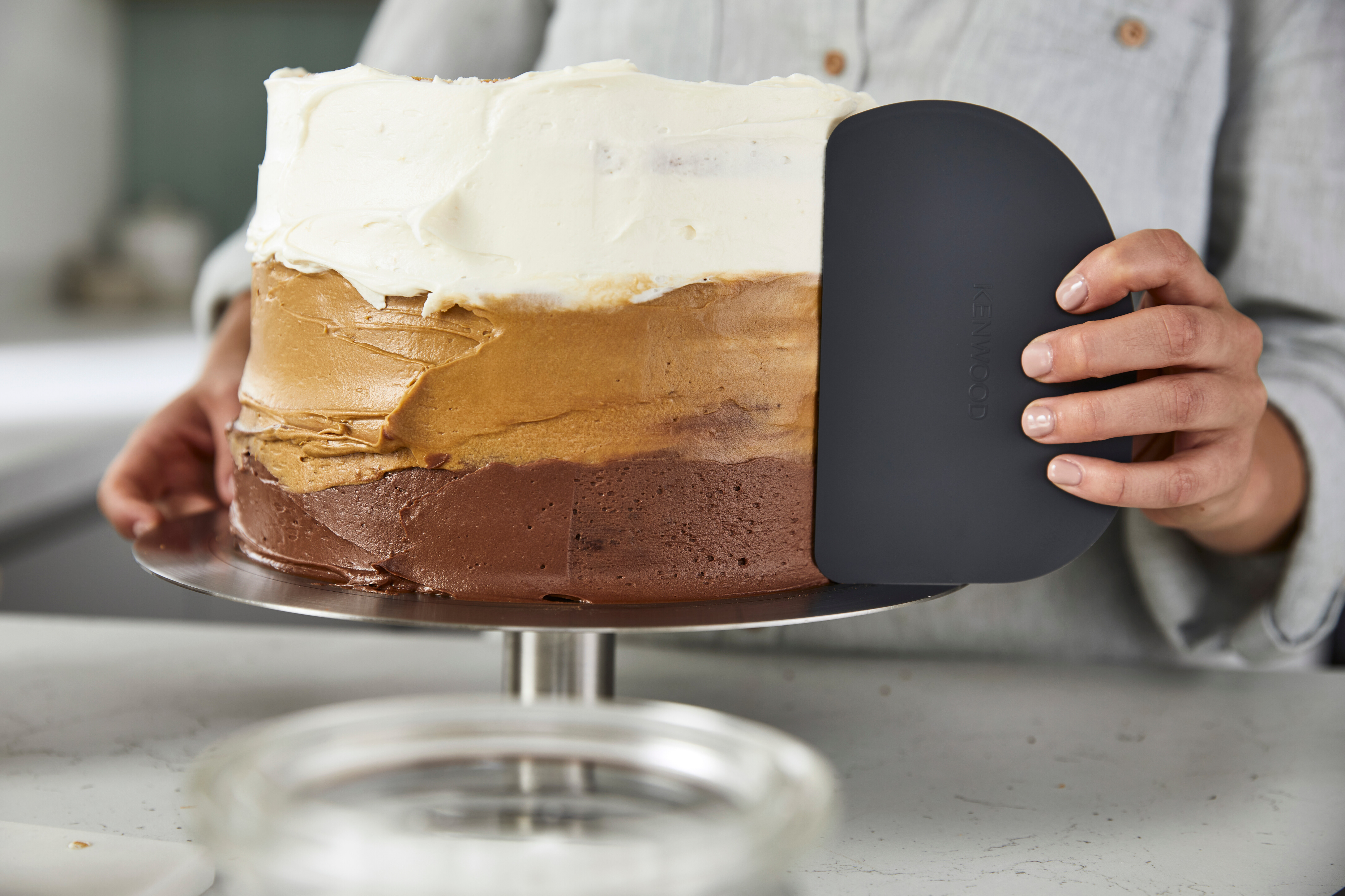 Ein mehrschichtiger Kuchen mit weißem, braunem und Schokoladenfrosting wird mit einem Schaber glattgezogen.