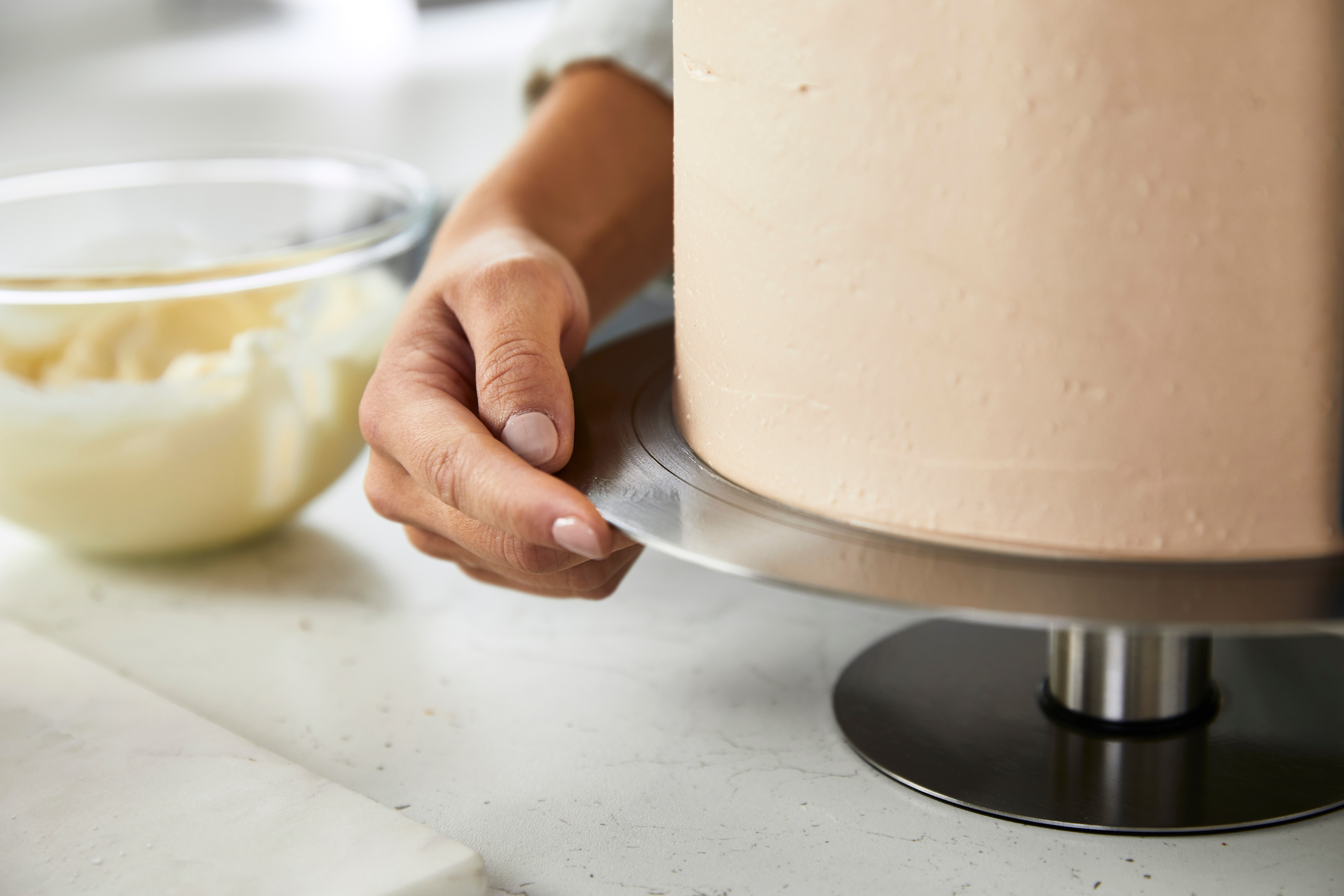 Eine Person dekoriert einen Kuchen mit Zuckerguss auf einem Kuchenständer. Eine Schüssel Zuckerguss ist im Hintergrund.