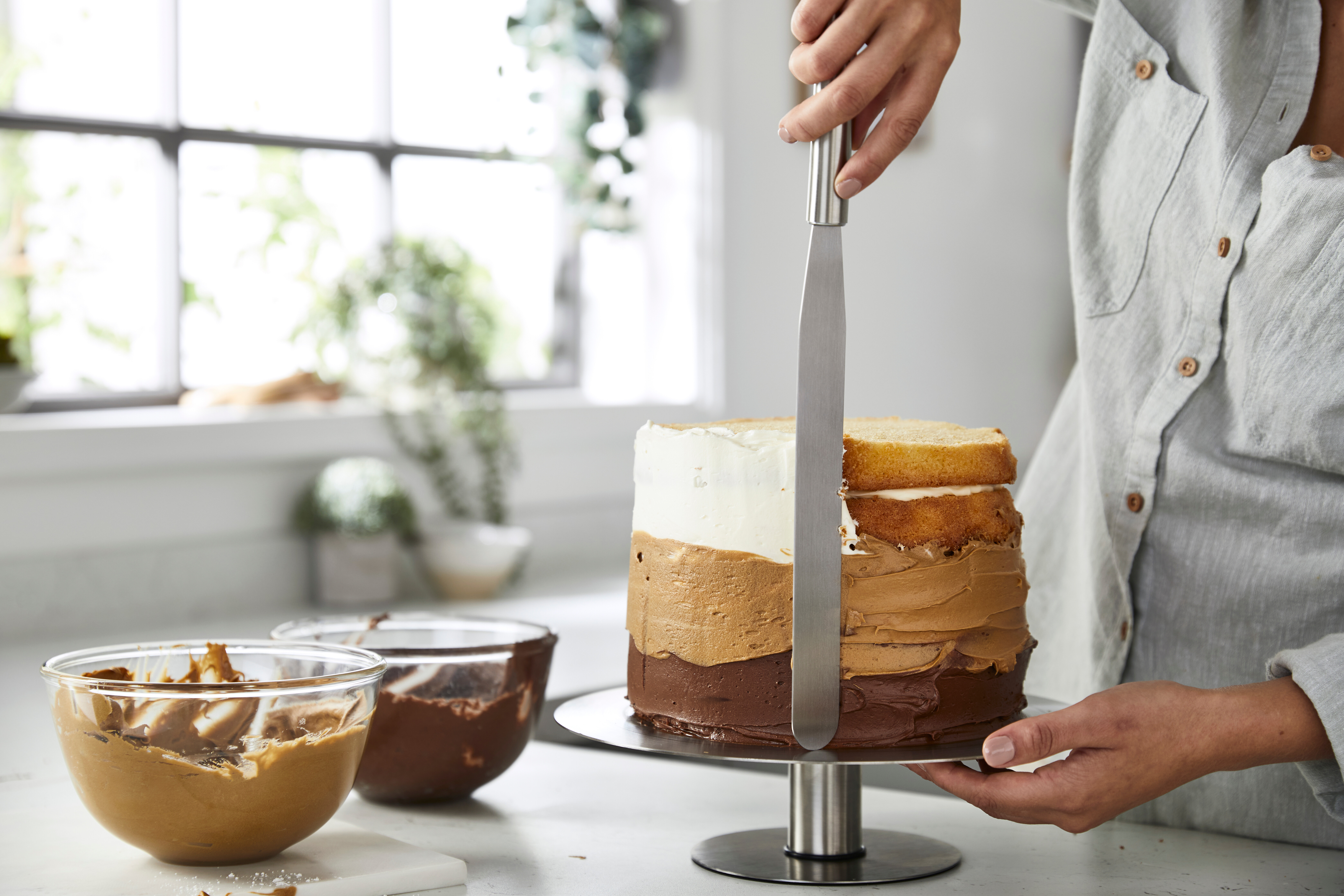 Eine Person, die einen geschichteten Kuchen mit einem Spatel mit Creme bedeckt. Schüsseln mit Creme sind im Vordergrund.