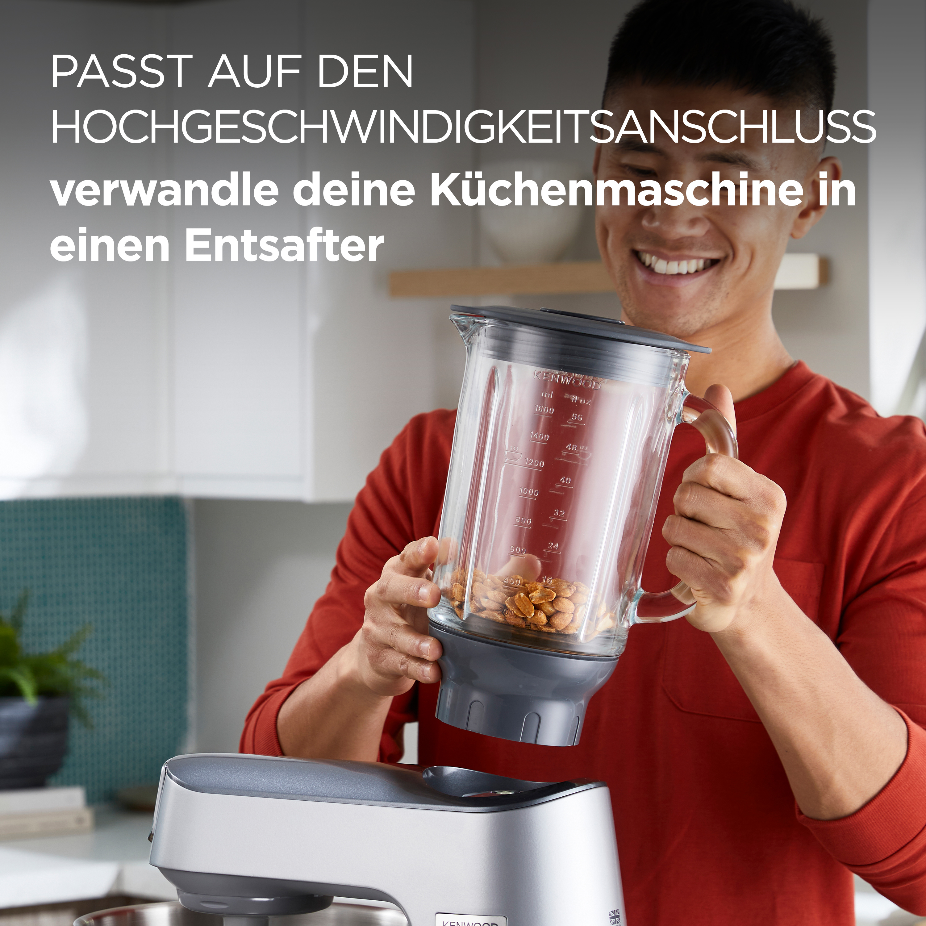 Ein lächelnder Mann hält einen durchsichtigen Mixerbehälter mit Nüssen neben einer silbernen Küchenmaschine.