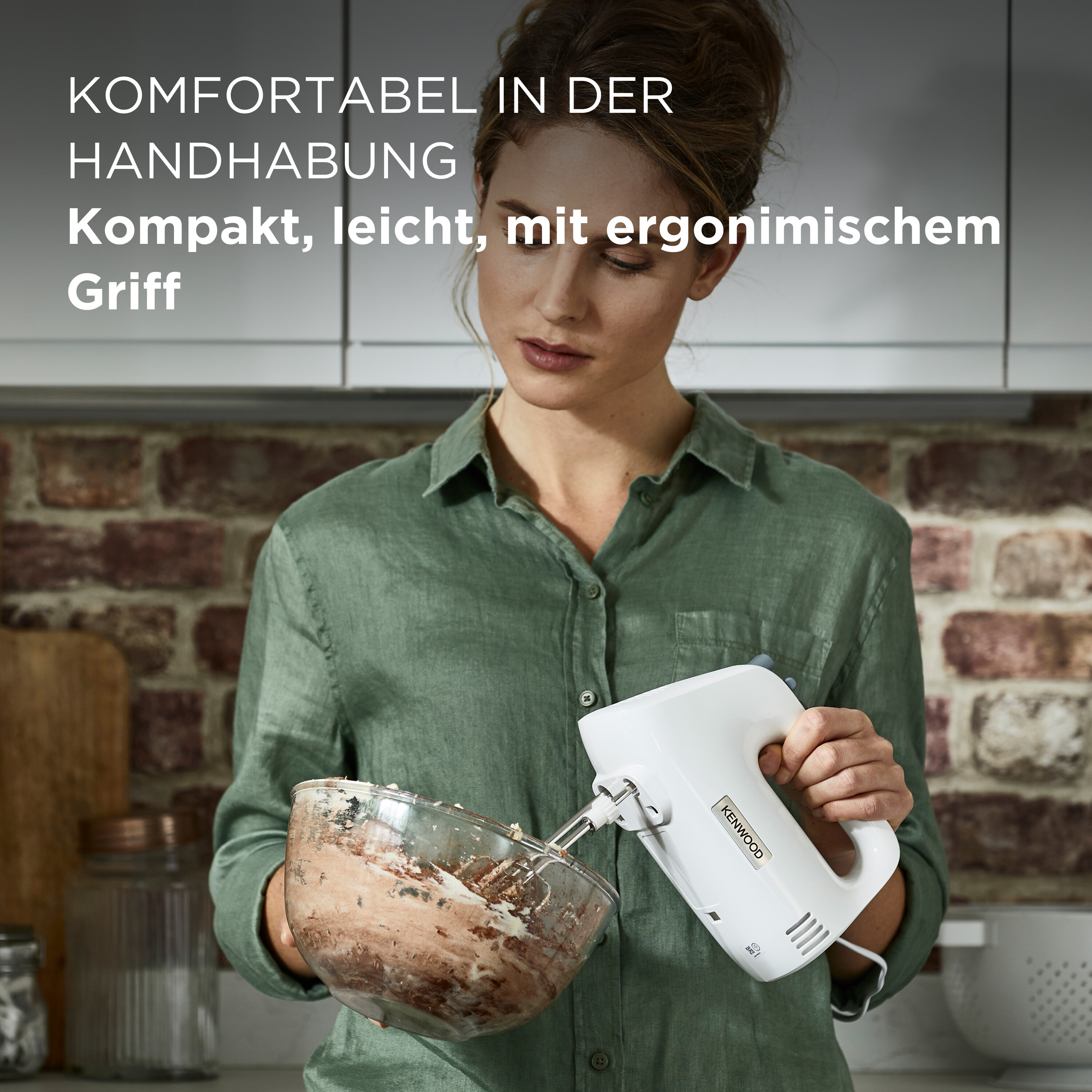 Frau hält weißen Kenwood-Mixer über Schüssel mit Teig. Hintergrund Backsteinmauer.