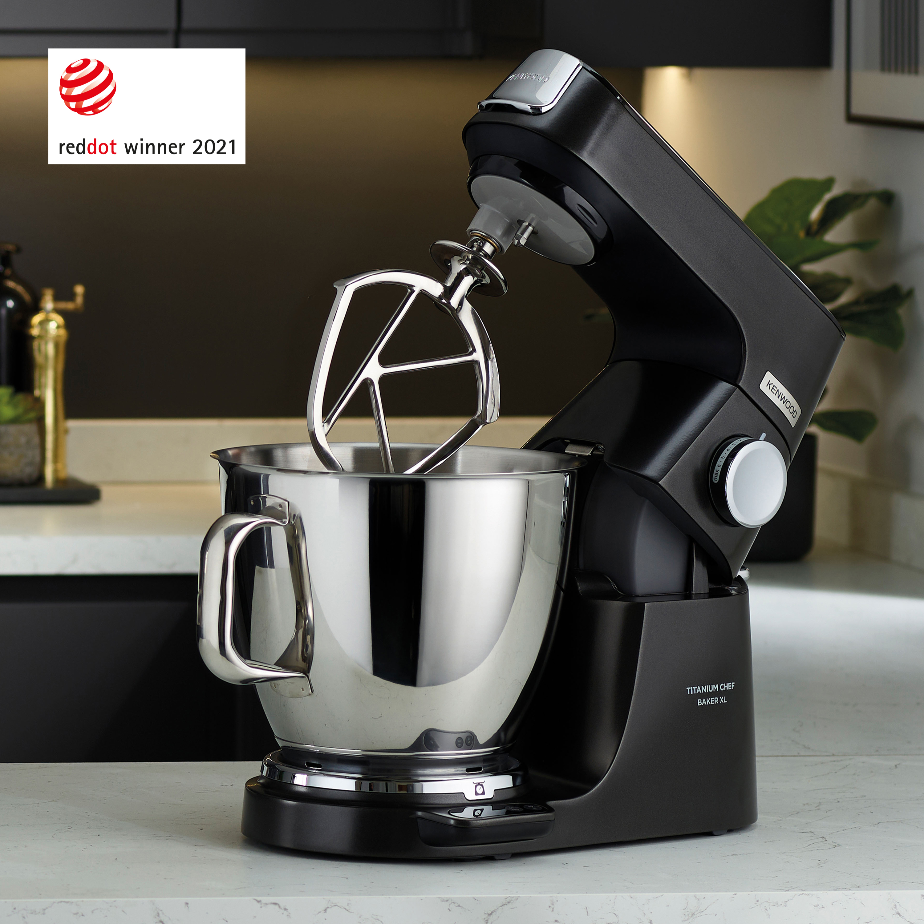 Schwarzer Küchenmixer mit silberner Schüssel auf weißer Arbeitsplatte. Reddot-Logo.