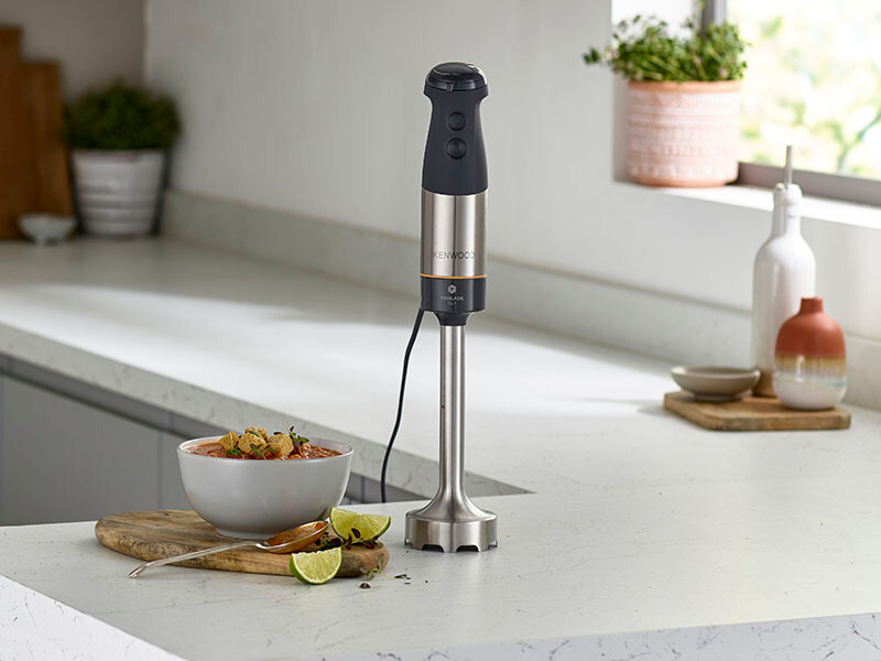 Ein silberner Stabmixer mit einer Schüssel Essen, Gemüse und Limette auf Theke.