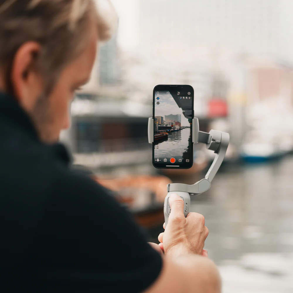 Ein Mann filmt mit einem Telefon auf einem Gimbal. Der Bildschirm zeigt Wasser und Gebäude.