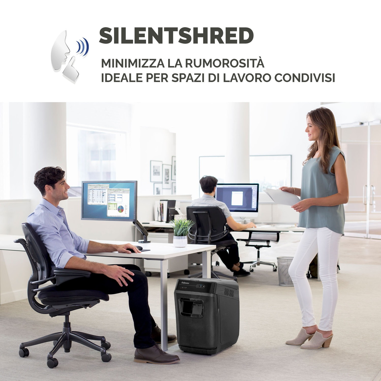 Scena d'ufficio. Un uomo alla scrivania, una donna con documenti, un tritadocumenti e due monitor.