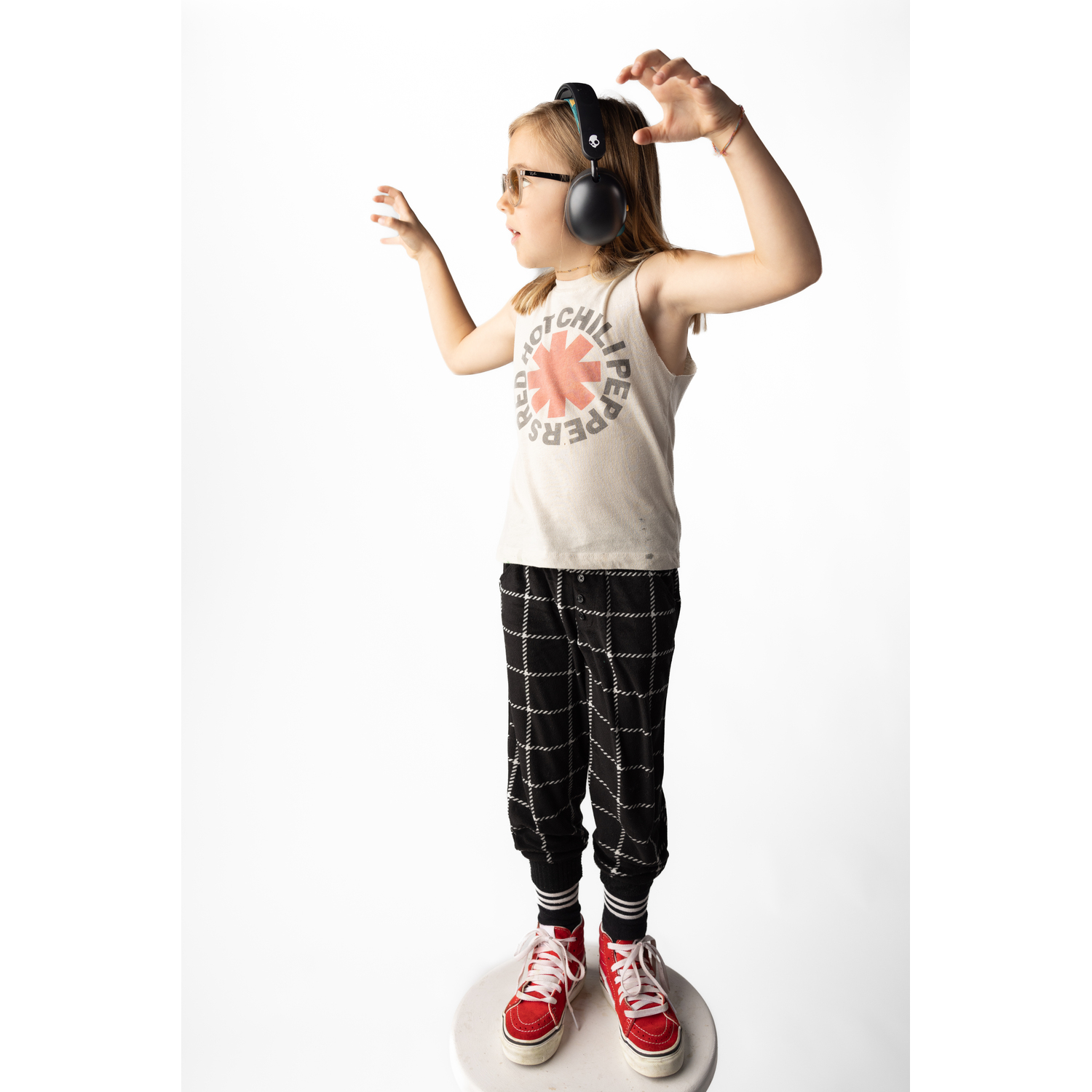 A young girl wearing headphones, glasses, and a Red Hot Chili Peppers shirt stands on a stool.