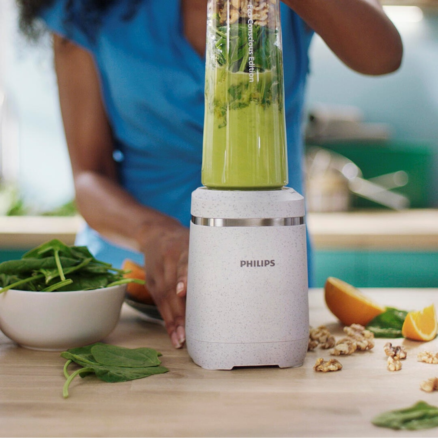 Persona che frulla ingredienti per frullato verde in un frullatore Philips bianco e grigio. Tavolo in legno.