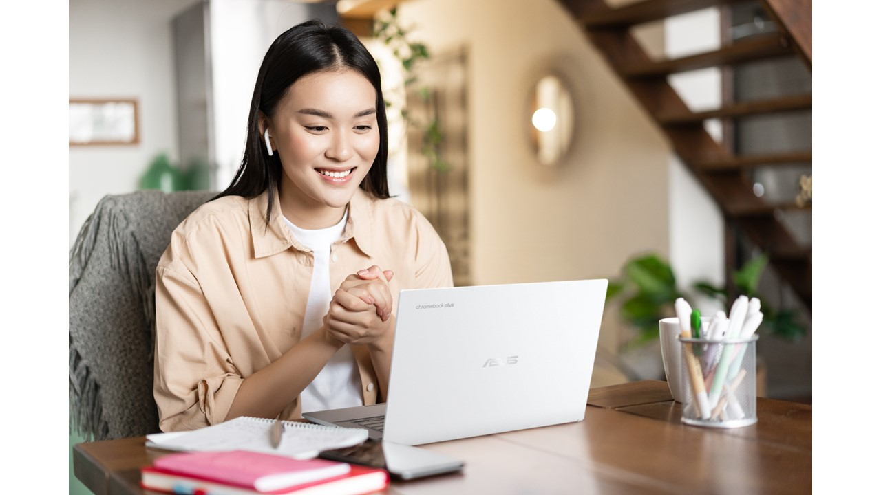 Eine Frau beim Videokonferieren auf einem weißen Laptop, lächelnd, mit Tisch und Treppe im Hintergrund.