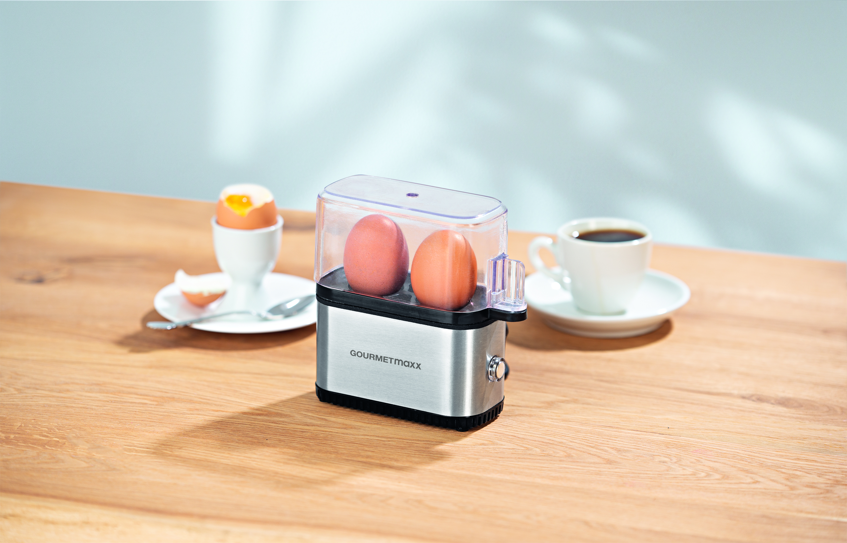 Eierkocher mit zwei Eiern, neben einer Tasse Kaffee und einem weichgekochten Ei.