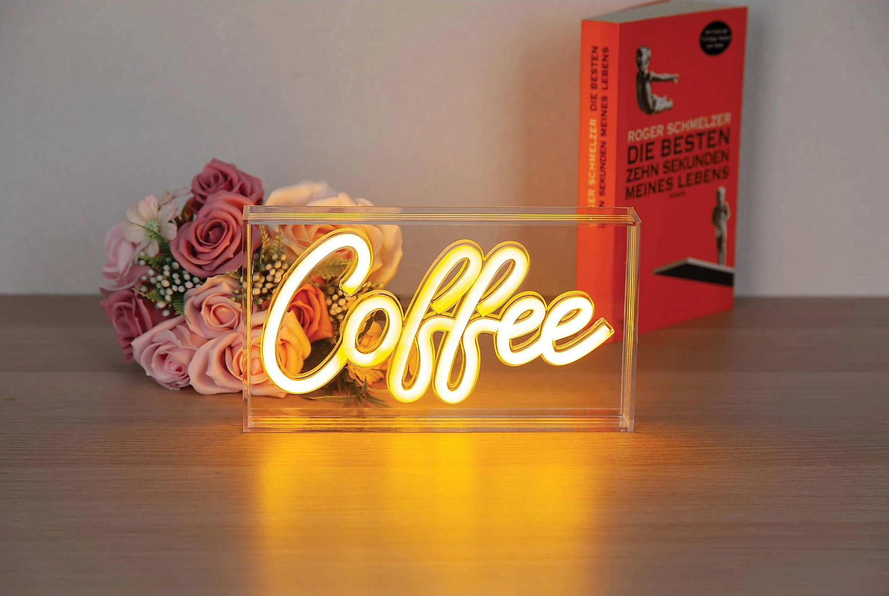 Neonowy szyld z kawą jest w przezroczystym pudełku. Za nim bukiet i książka.