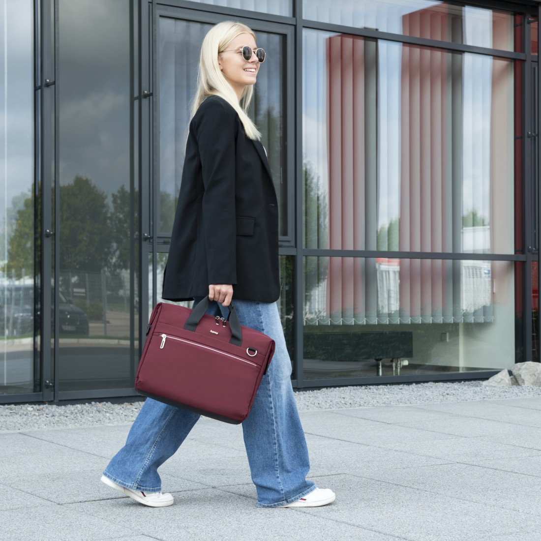 Eine Frau geht mit burgunderfarbener Laptoptasche in der Nähe eines Glasgebäudes.