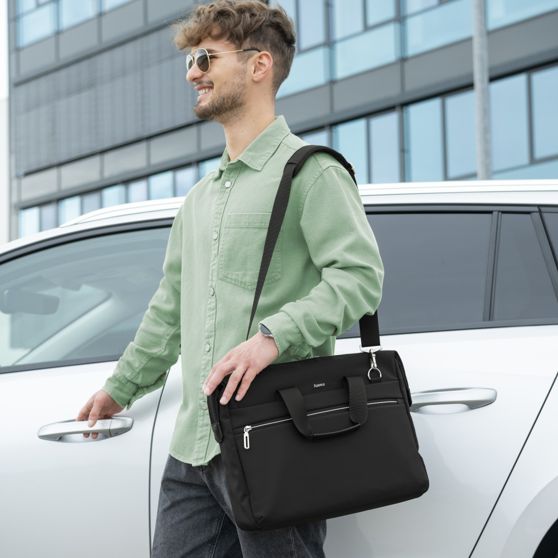 Ein Mann in grünem Hemd und Sonnenbrille trägt eine schwarze Tasche neben einem weißen Auto.
