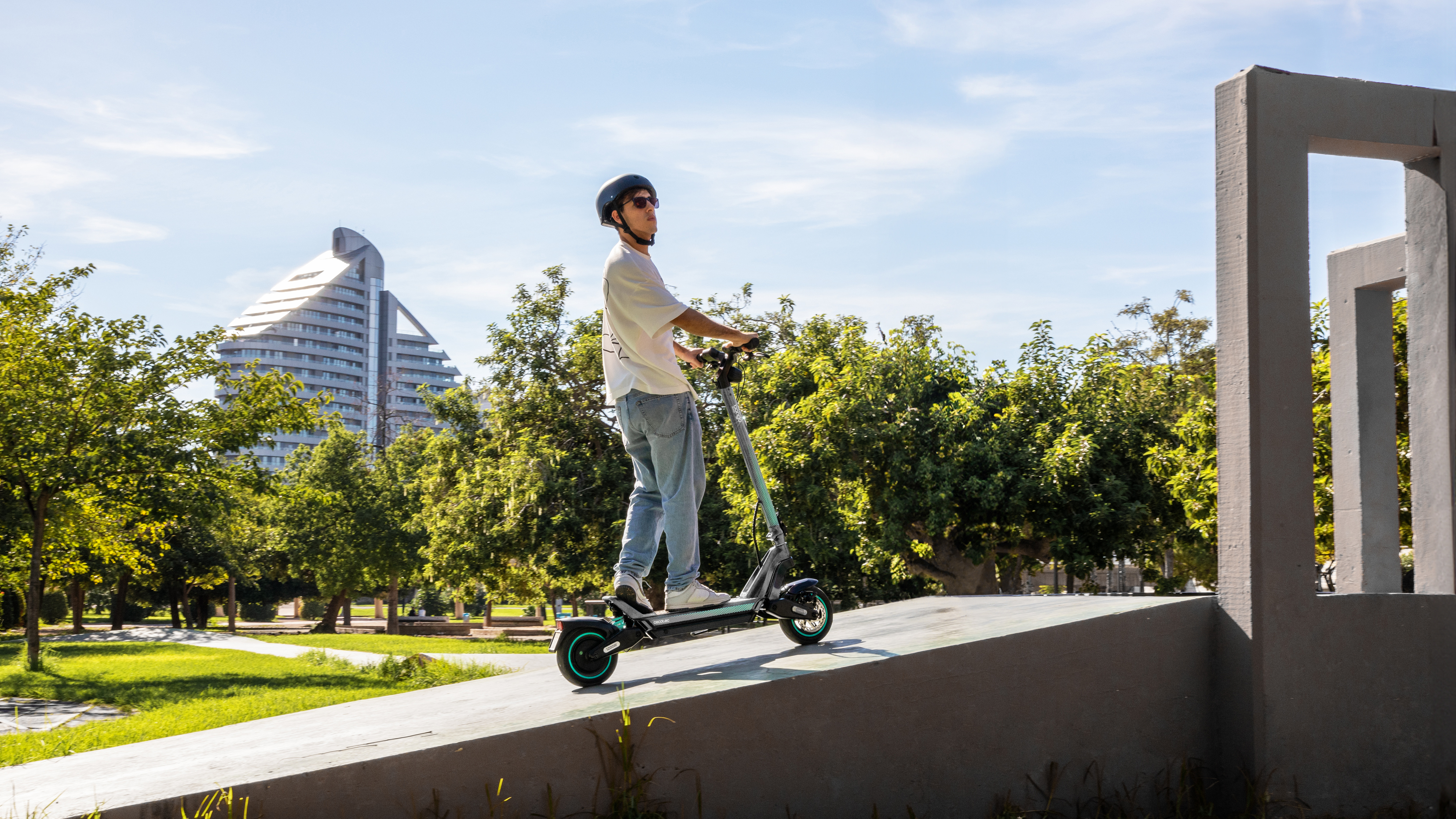 Un hombre montando un patinete en una rampa, edificios de la ciudad y árboles al fondo. Día soleado.
