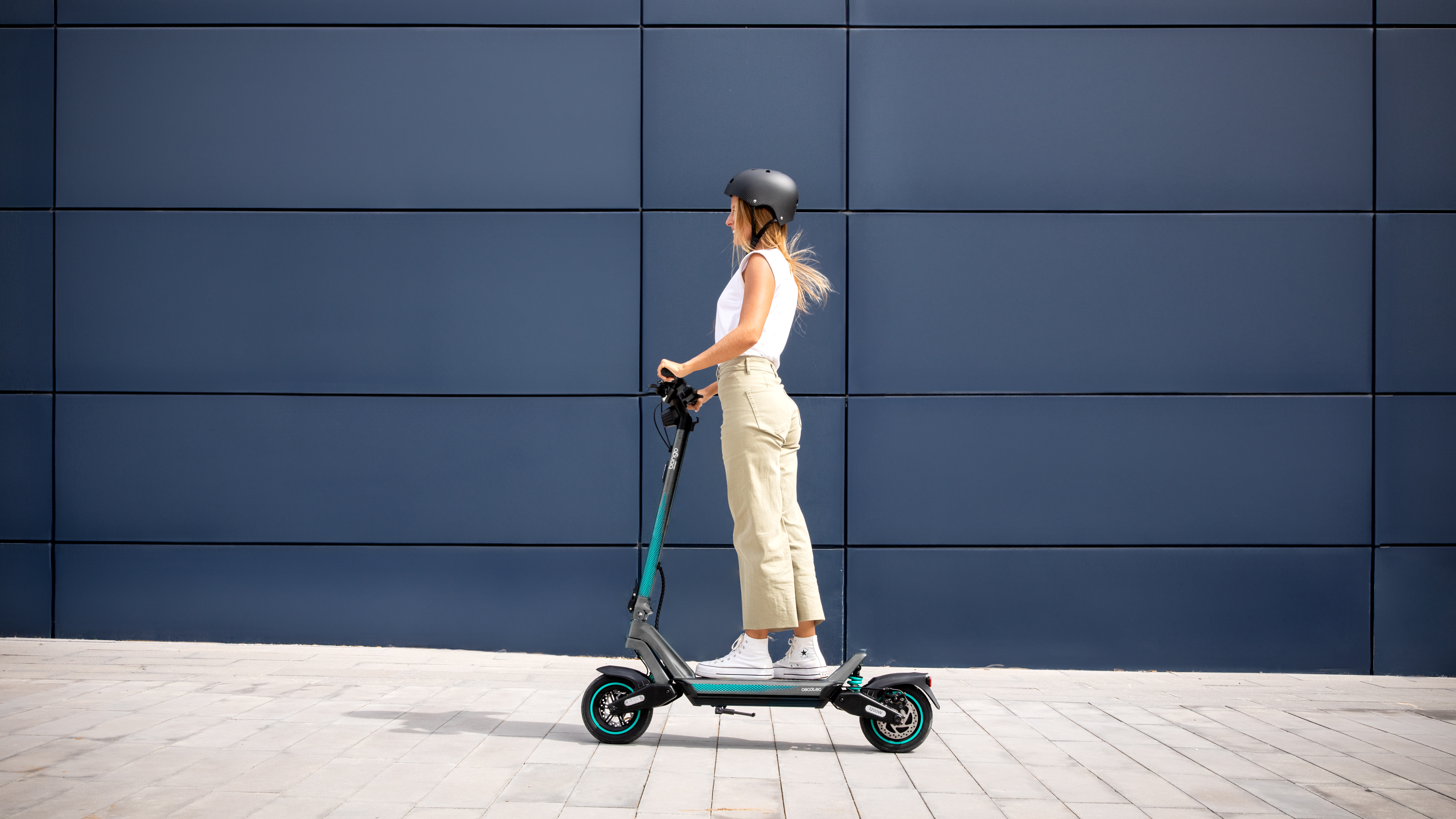 Una mujer montando un patinete eléctrico turquesa y negro en una acera.