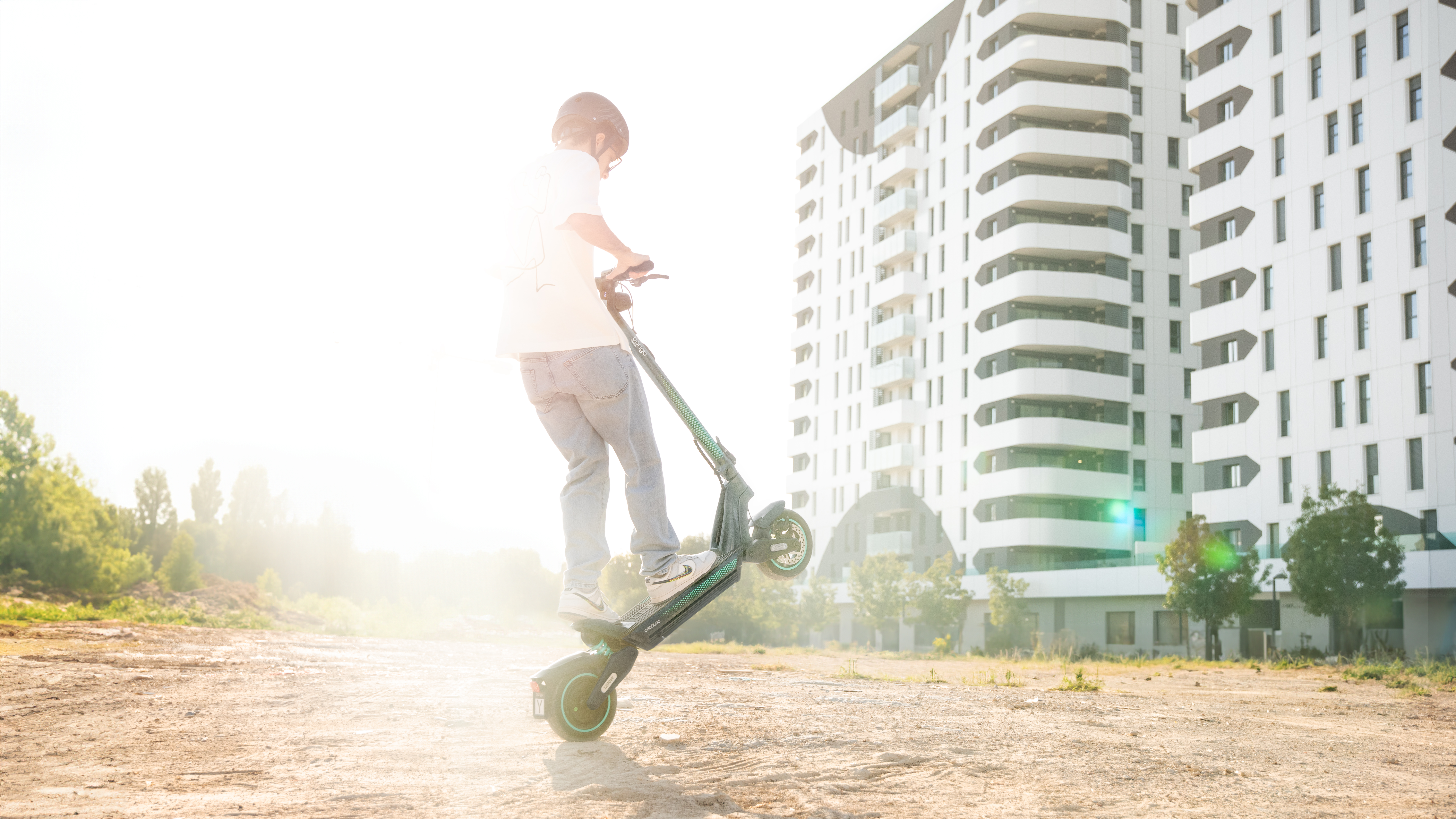 Una persona montando un patinete eléctrico haciendo un caballito. Un edificio alto al fondo.