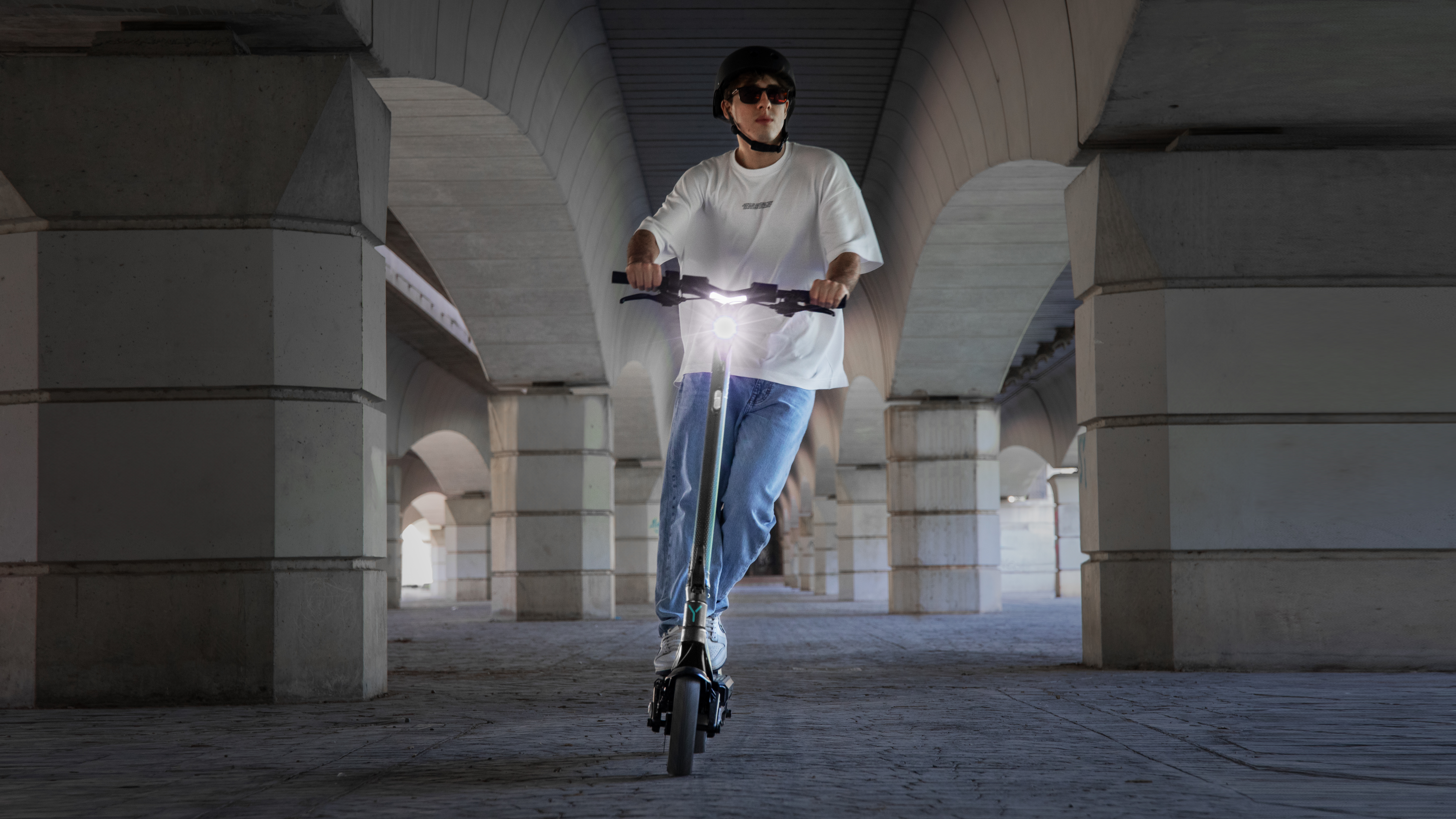 Una persona monta un patinete eléctrico bajo un puente arqueado. Faro encendido.