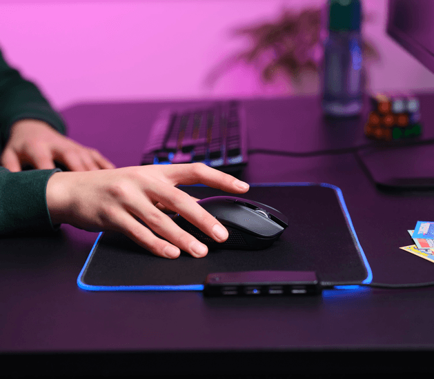 Személy kezében fekete gaming egér, fekete egérpadon, kék LED-es lámpákkal.