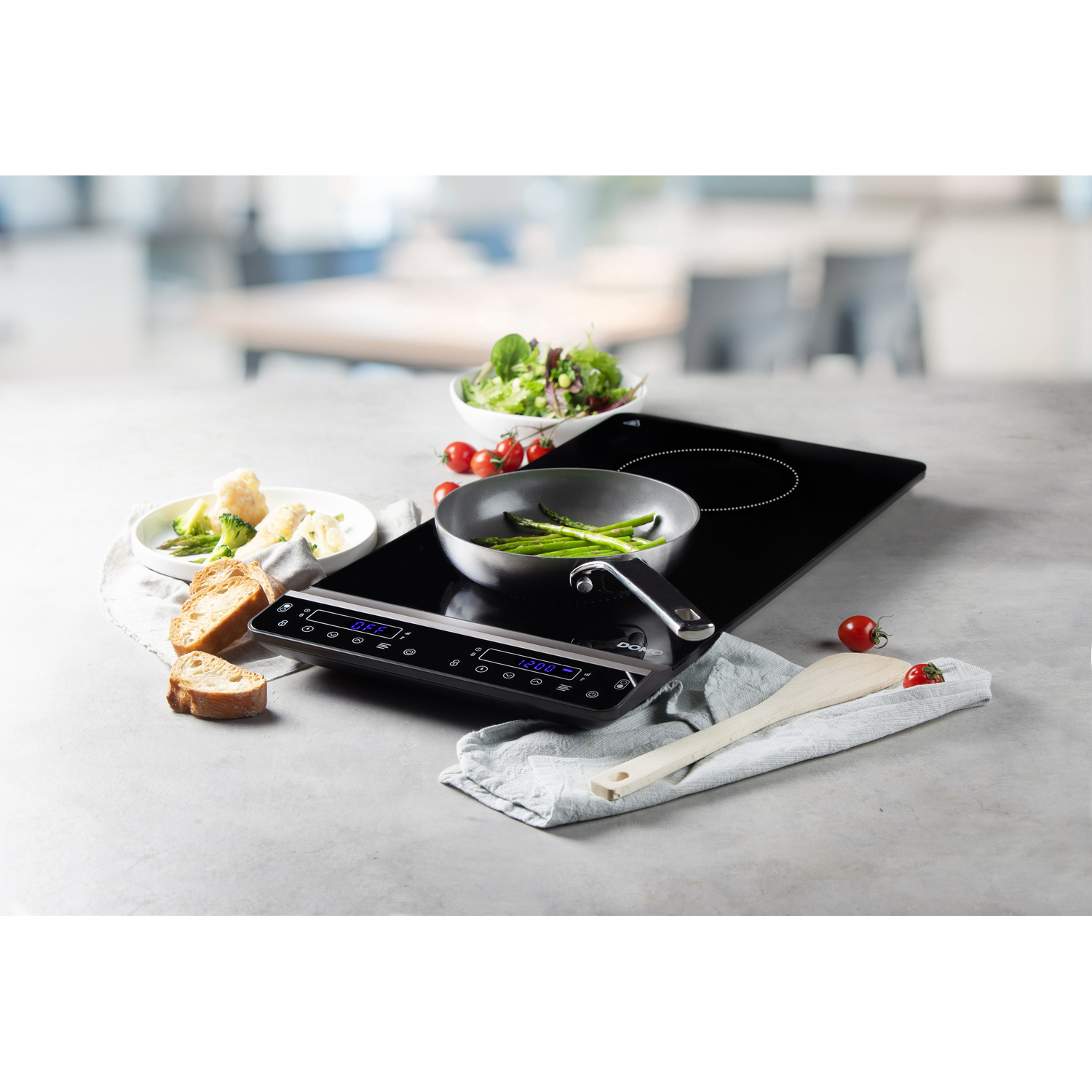 Black induction cooktop with food on it. Includes pan, asparagus, and bread. Wooden spoon on counter.