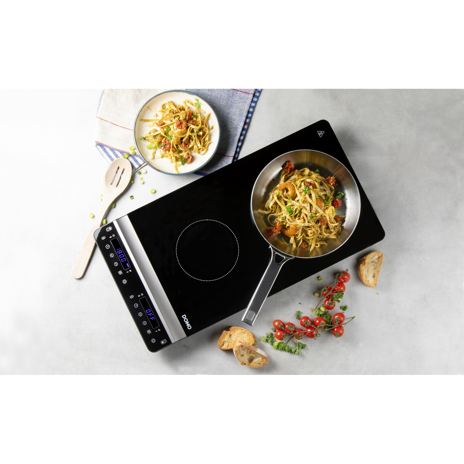 A black induction cooktop with a pan of pasta and a plate of pasta, tomatoes and bread.