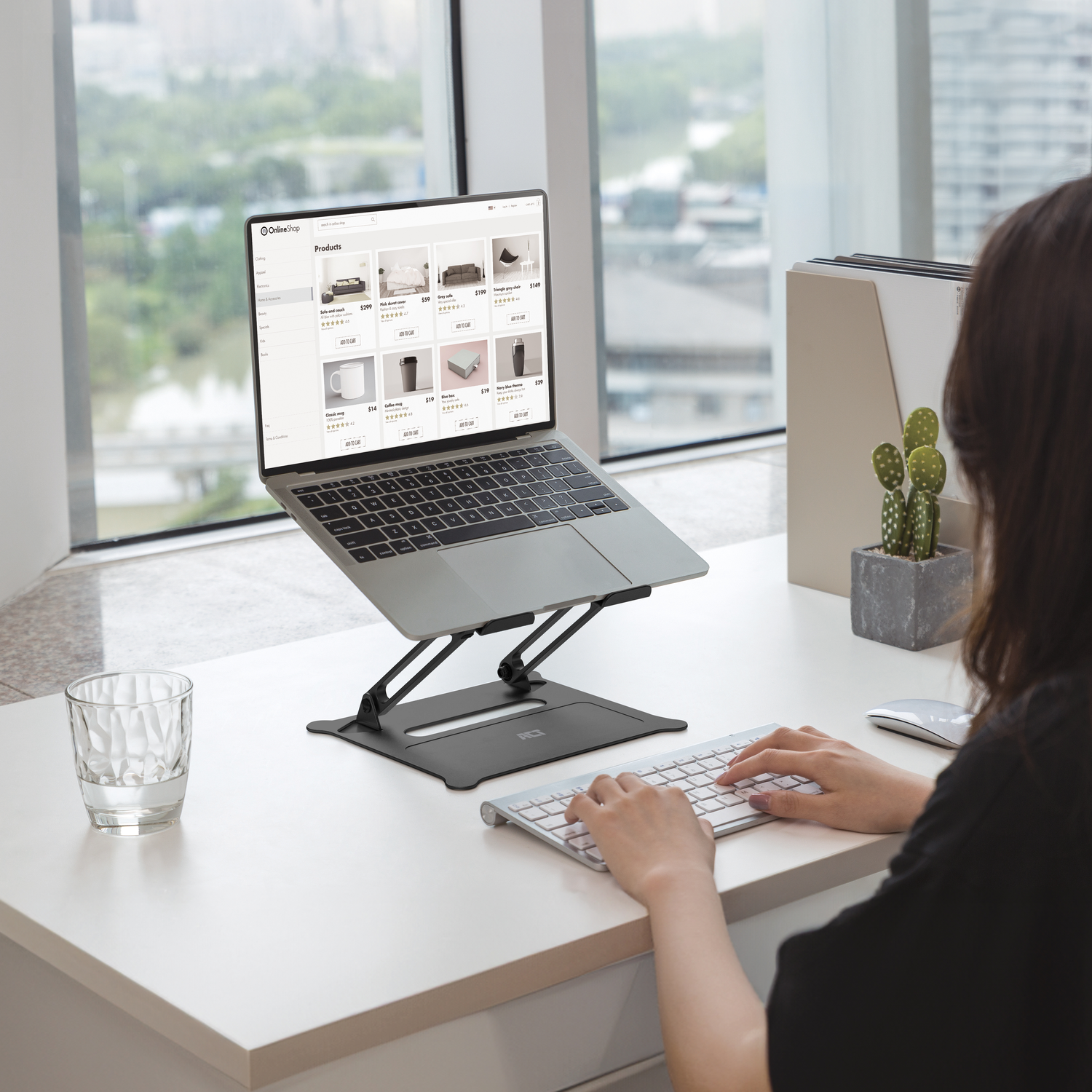Persoon die werkt op een laptop op een standaard. Raam op de achtergrond, kopje en plant.