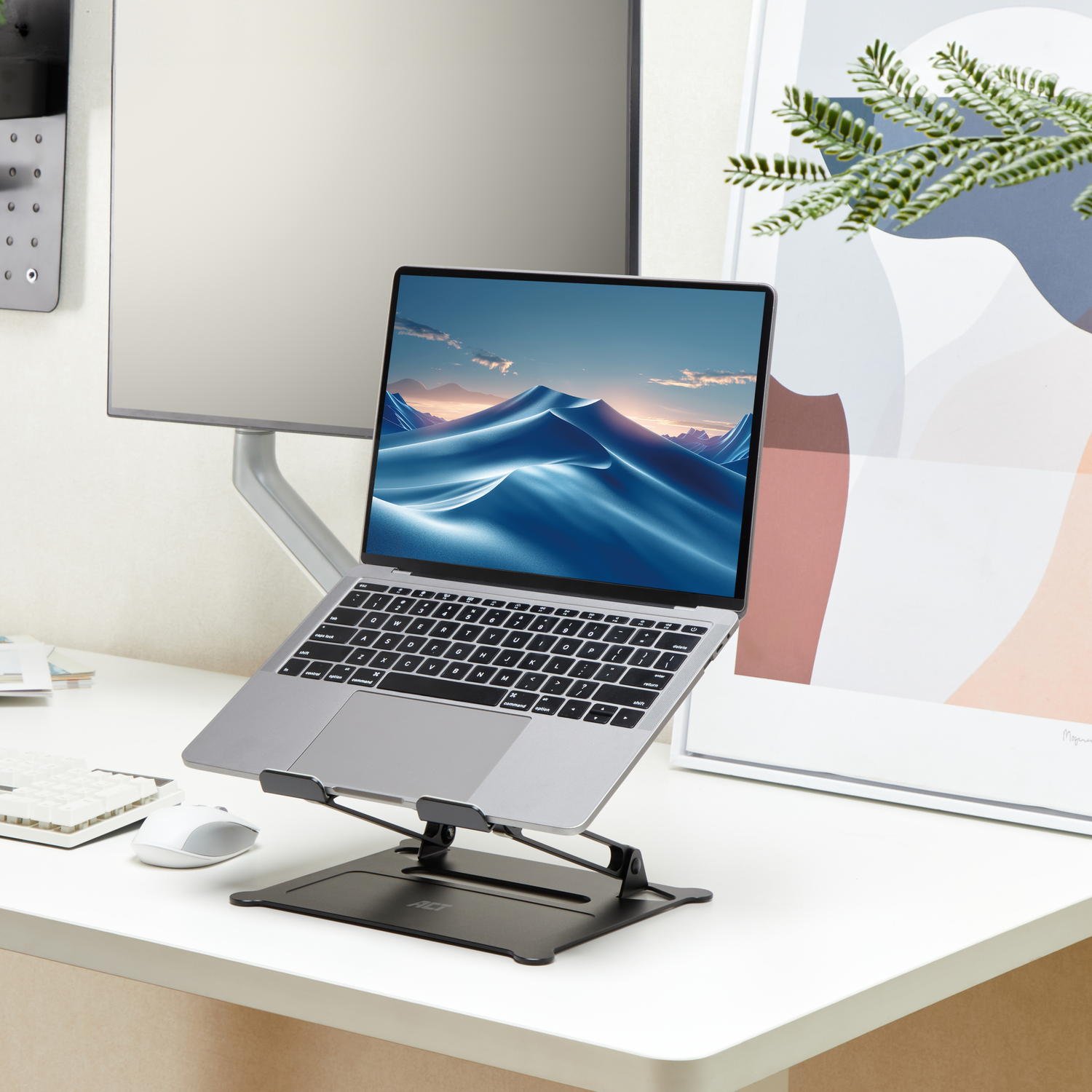 Een laptop op een standaard op een wit bureau, met een monitor en een kunstafdruk.