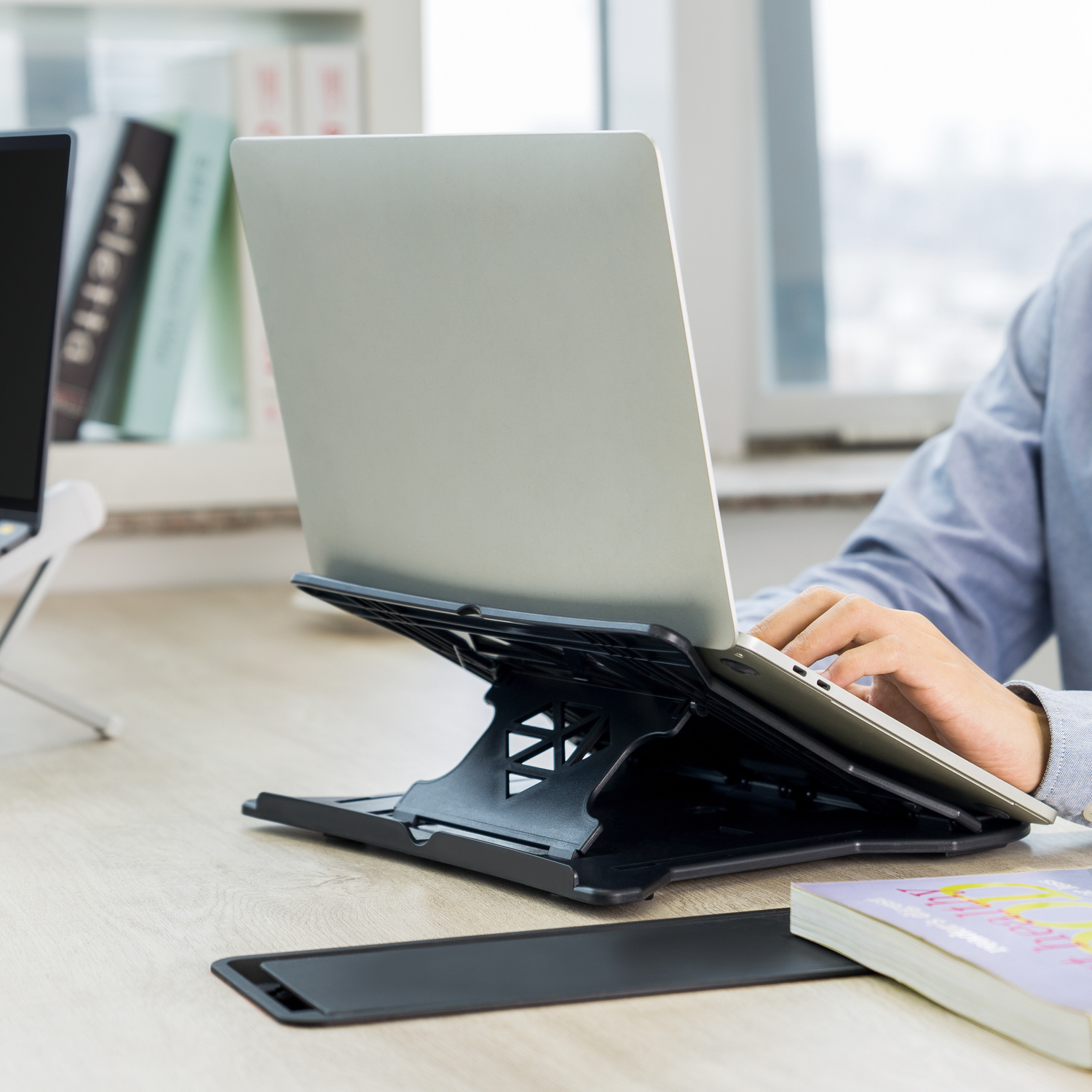 A person using a laptop on a stand. A book and a tablet are on the desk.
