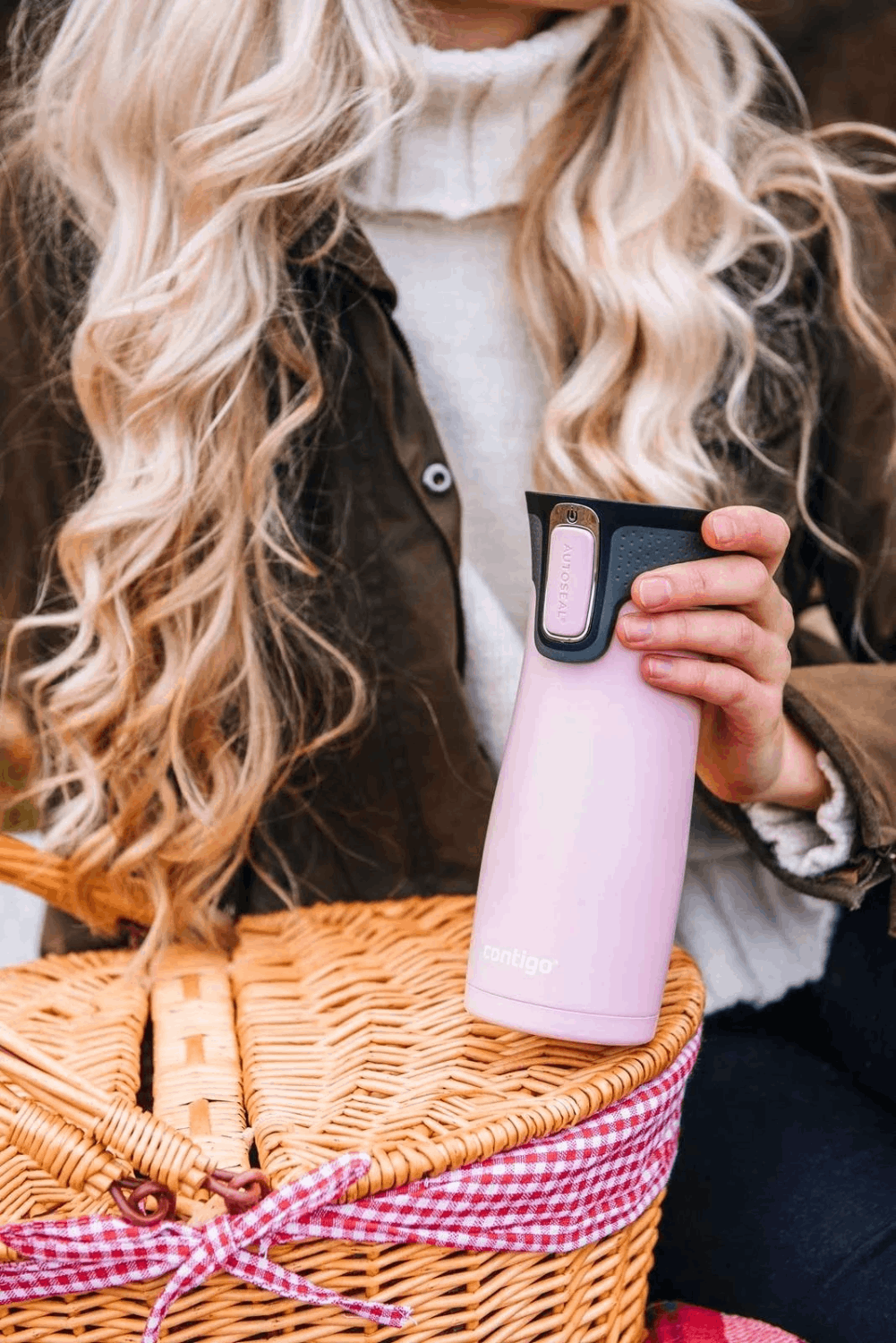 Una mujer sostiene un termo Contigo rosa junto a una cesta de picnic. Cabello rubio, desenfocado.