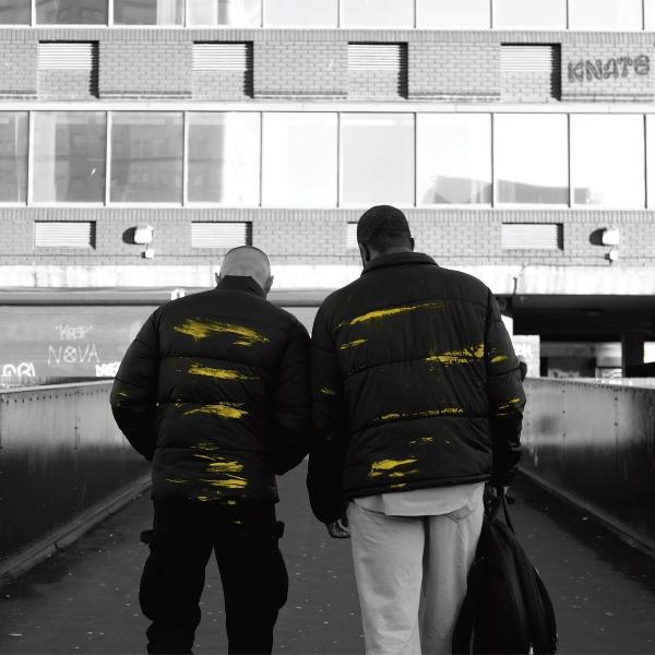 Zwei Männer gehen weg auf einer Brücke. Sie tragen schwarze Jacken mit gelben Farbstrichen.