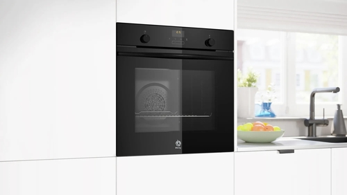 Un horno empotrado en un armario de cocina blanco. Hay una ventana y un cuenco de fruta en el fondo.