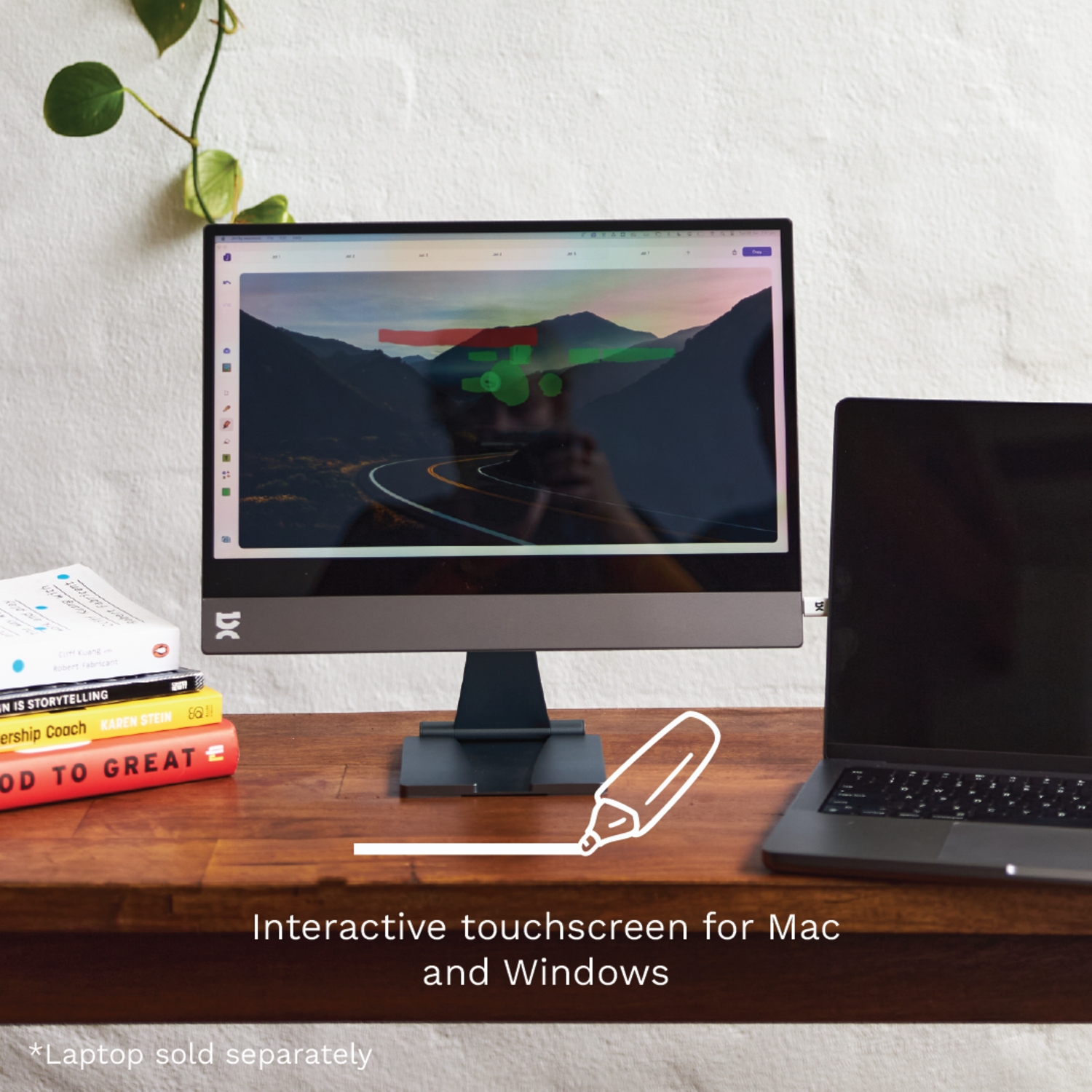Interactive touchscreen monitor with a laptop and books on a wooden desk. Green plant.