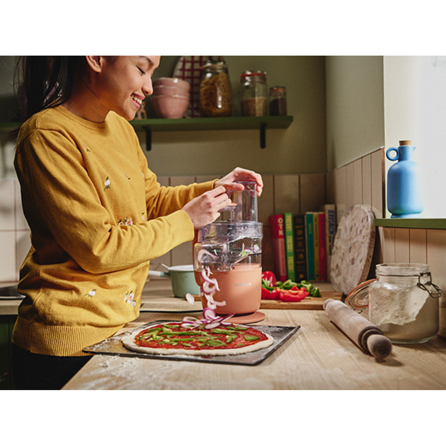 Femme en jaune cuisine. Elle utilise un robot pour préparer les ingrédients de la pizza.