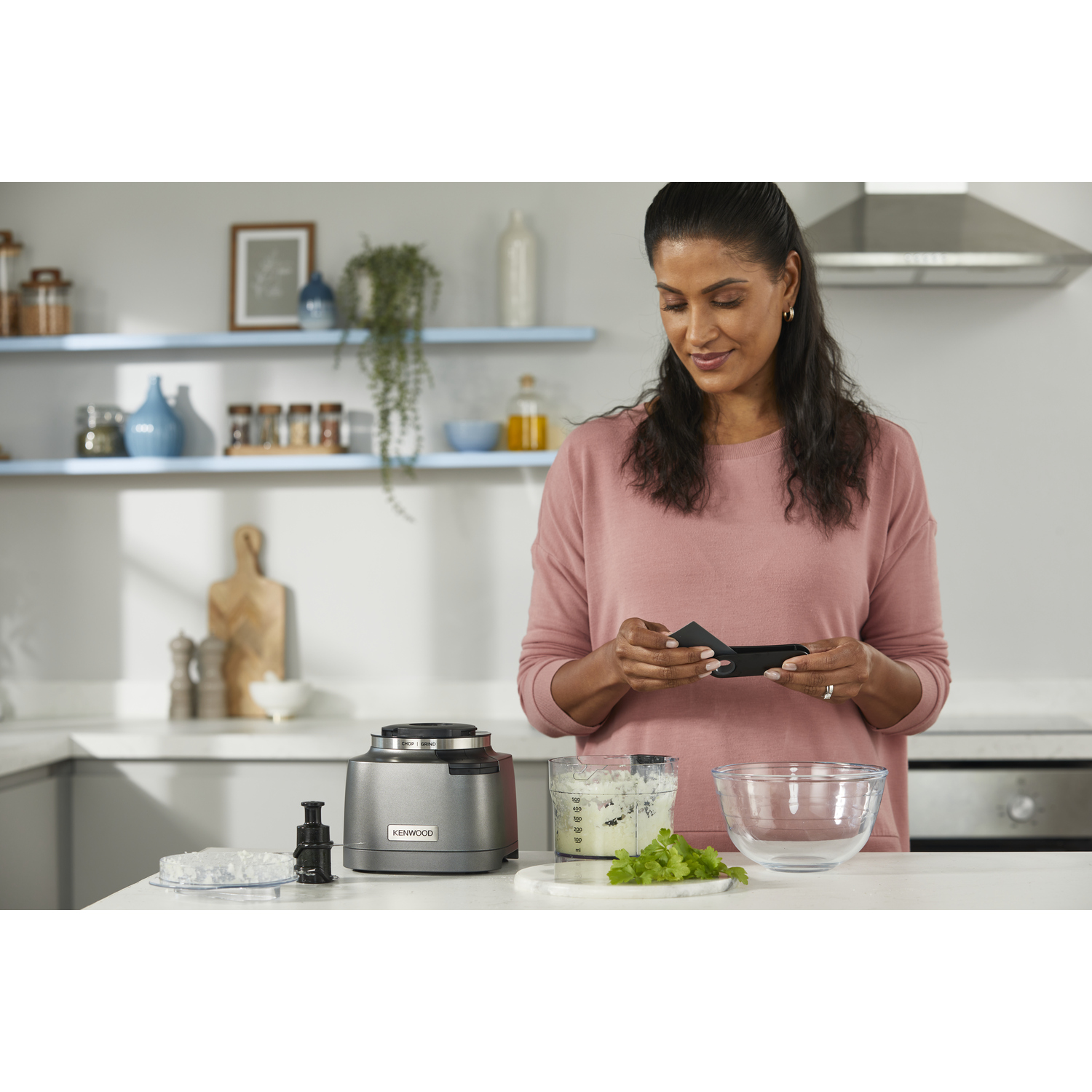 Vrouw in keuken met keukenmachine, houdt zwart accessoire vast, lacht.