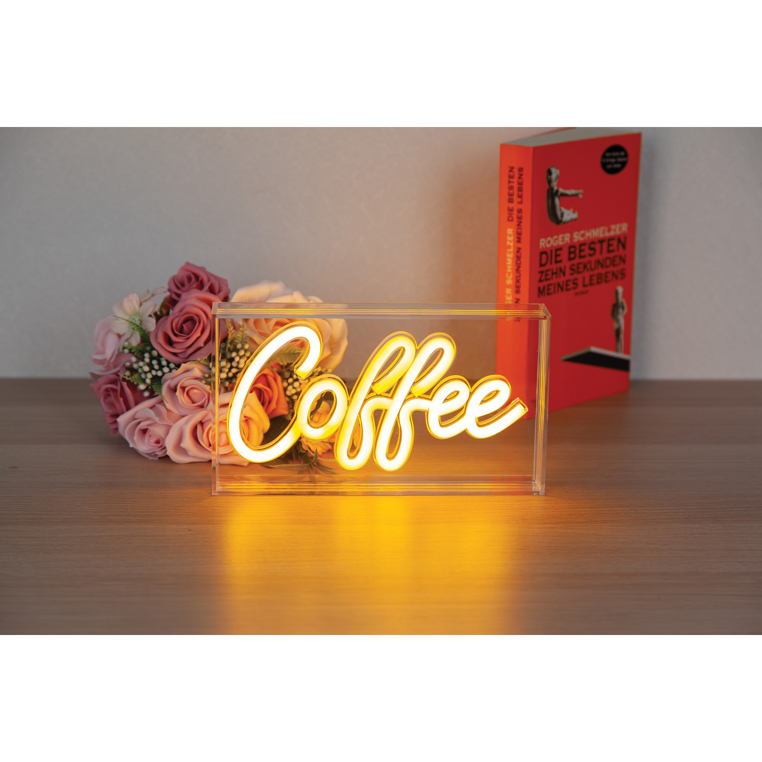A neon sign that says 'Coffee' on a table with flowers and a book.