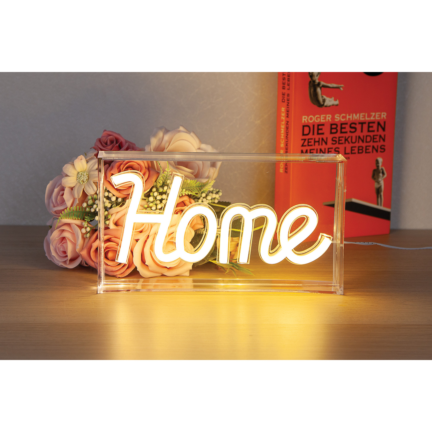 Glowing neon 'Home' sign in a clear box, with flowers and a book in the background.