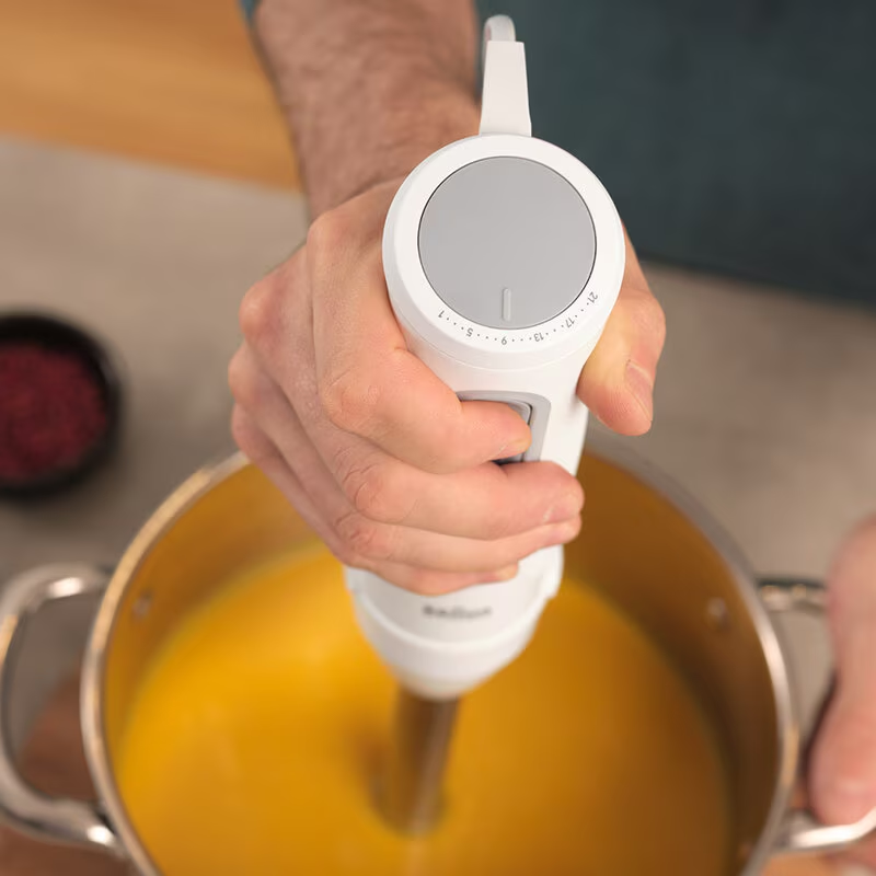 Mains d'une personne tenant un mixeur plongeant blanc au-dessus d'une marmite de soupe jaune, fond: planche à découper.