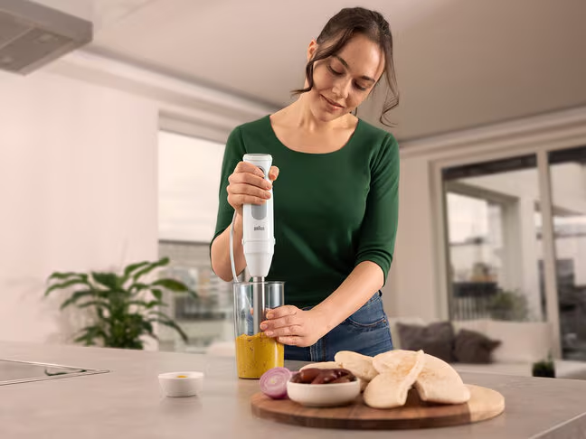 Femme utilisant un mixeur plongeant blanc dans une cuisine. Divers aliments sont visibles.