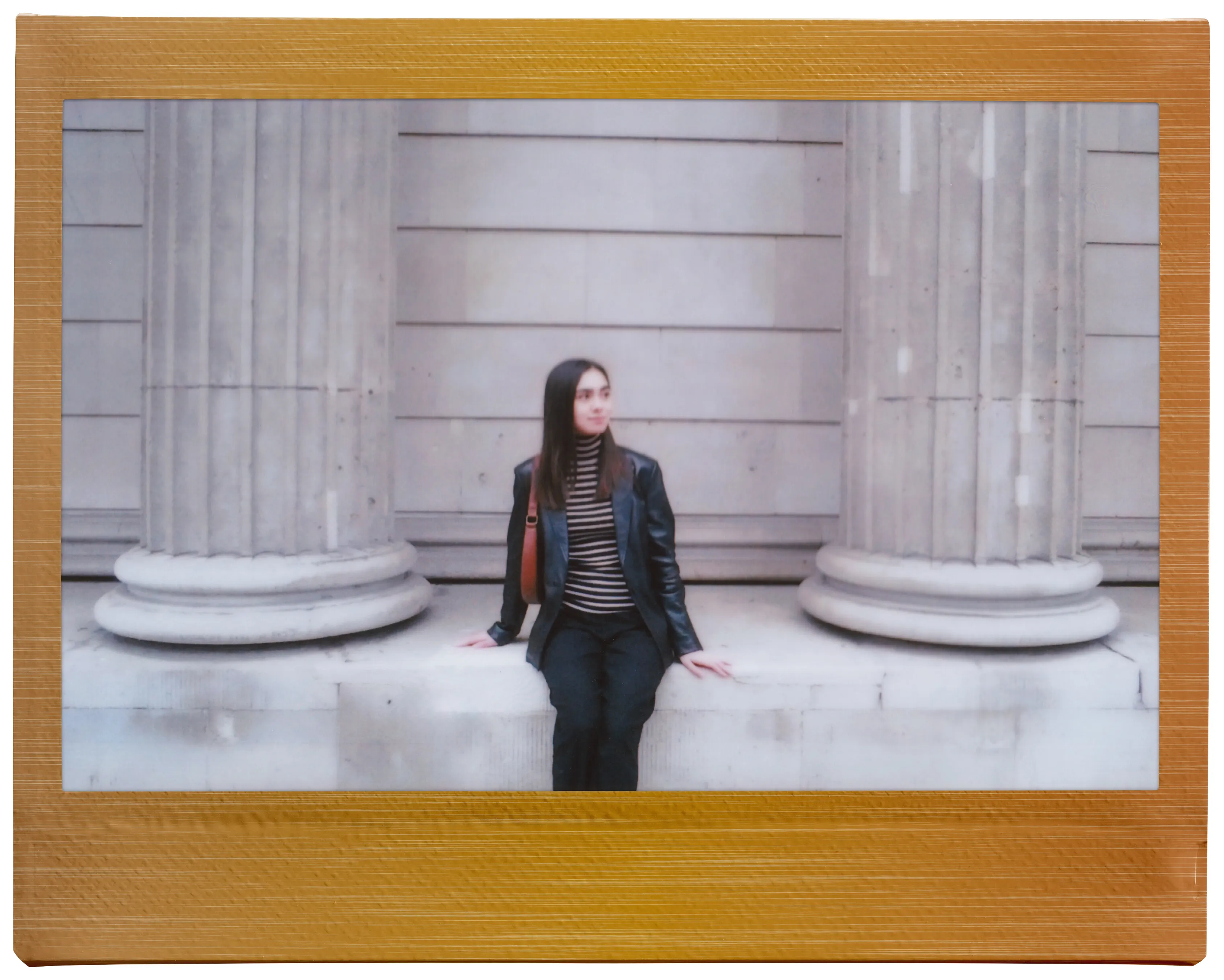 Femme assise sur un mur entre deux piliers. Porte une veste noire et une chemise rayée.