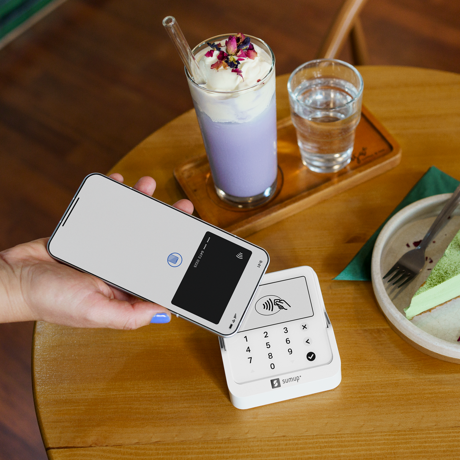 Hand hält Smartphone über ein Zahlungsterminal, daneben Getränke und Kuchen.