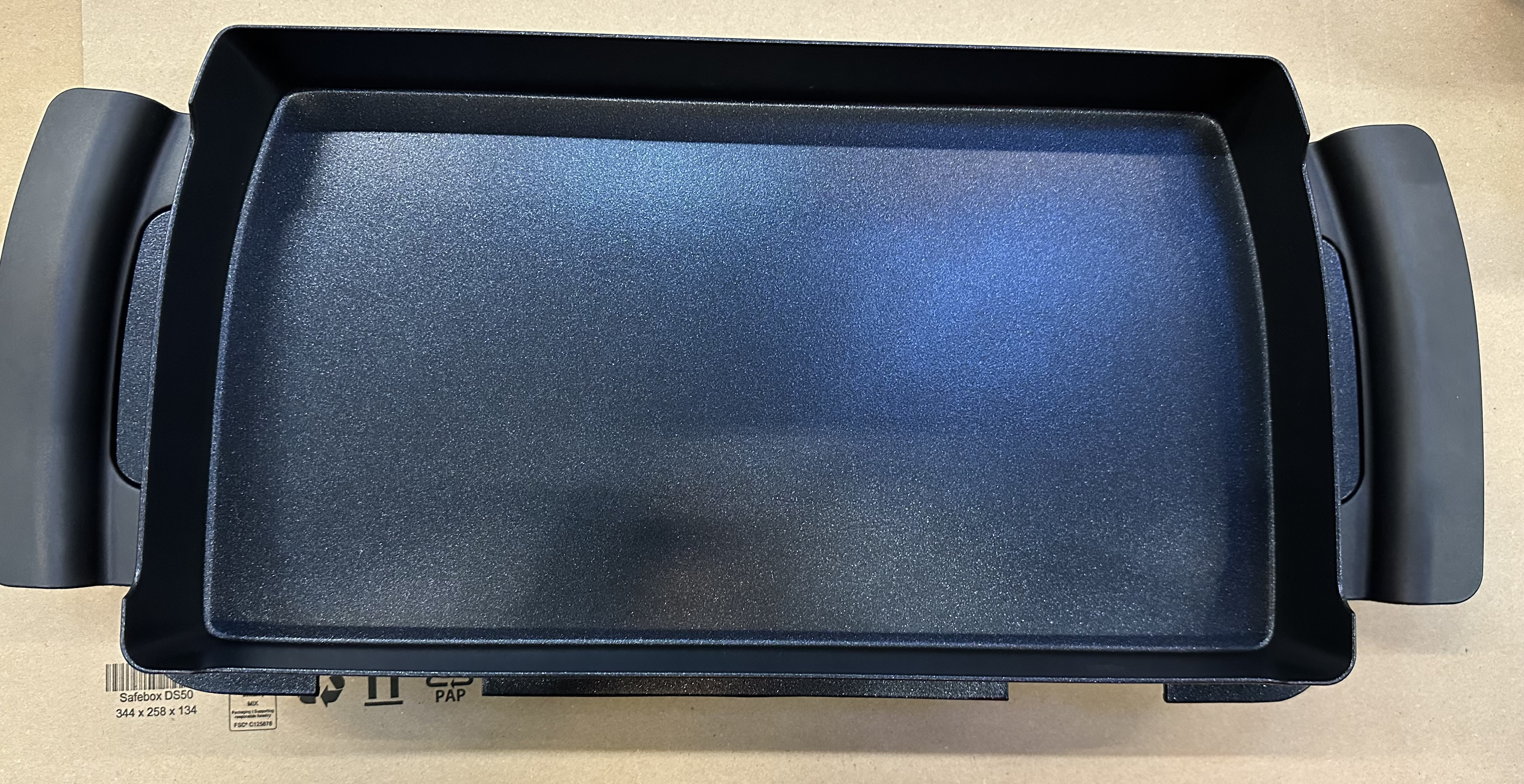 Czarna płyta grillowa z uchwytami, leżąca na kartonie.