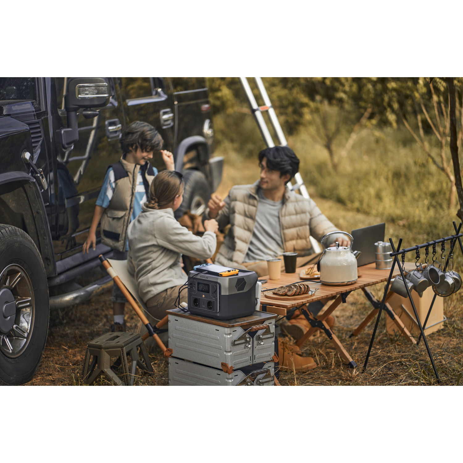 Family camping near a black SUV. They are seated at a table with a power station.